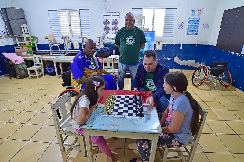 uas meninas jogam xadrez com auxílio de um instrutor. Ao fundo, dois homens observam, um deles em cadeira de rodas. A sala tem ambiente escolar e materiais pedagógicos visíveis.