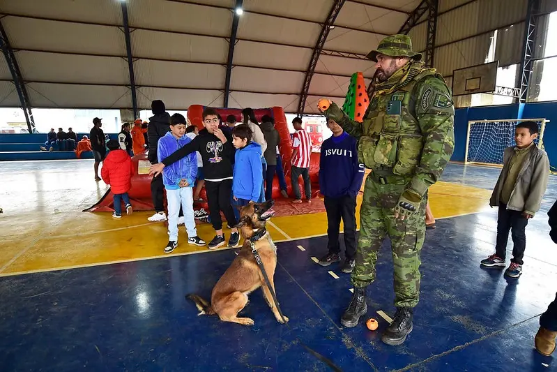 Crianças observam cão policial atento a comando de agente fardado que segura bolinha. Ao fundo, brinquedos infláveis e demais participantes acompanham a demonstração com interesse