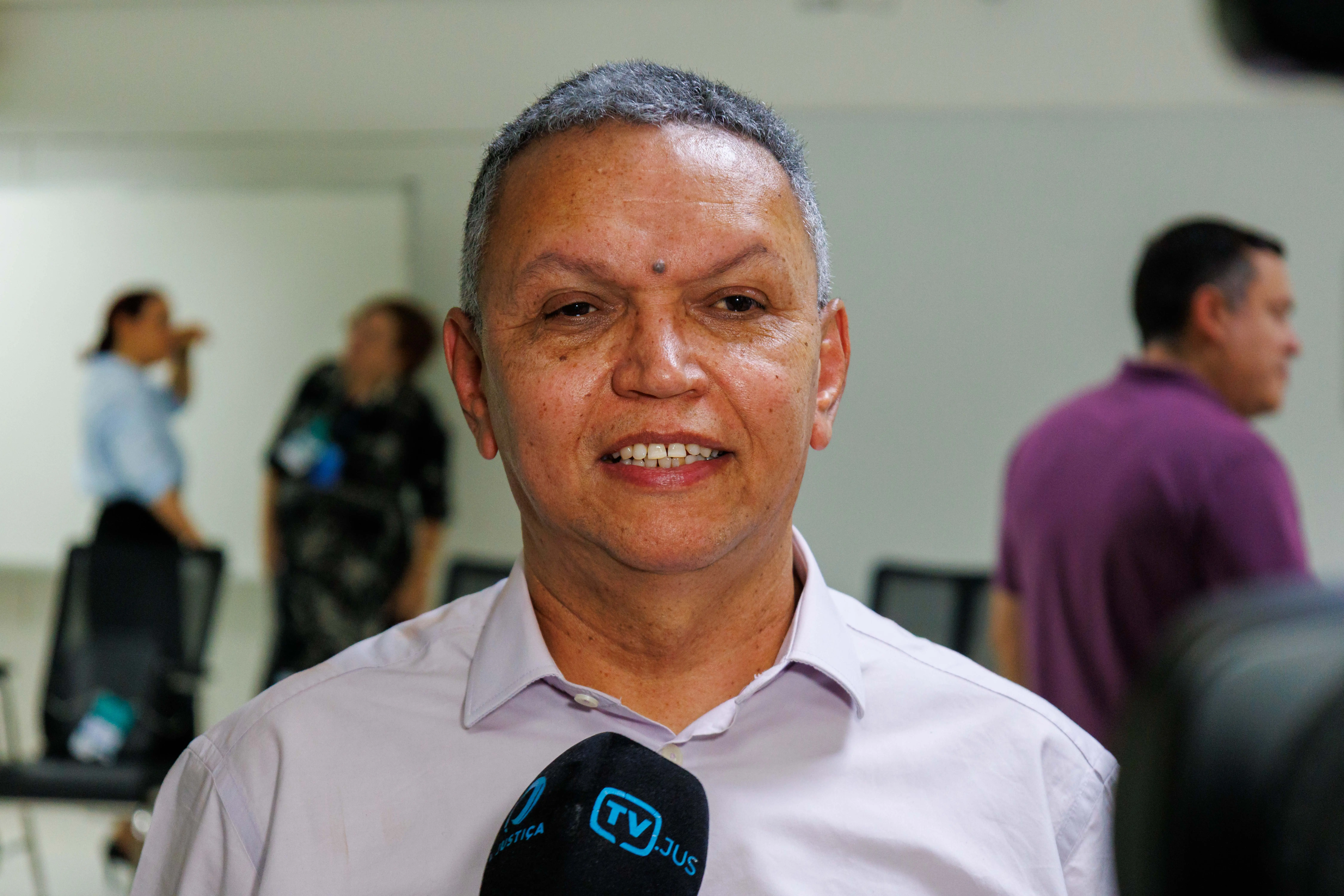 Servidor José Nivaldo usa camisa clara segura microfone da TV Justiça. Rostos desfocados. Ao fundo, outras pessoas em pé e sentadas. Ambiente interno, sugerindo evento ou entrevista