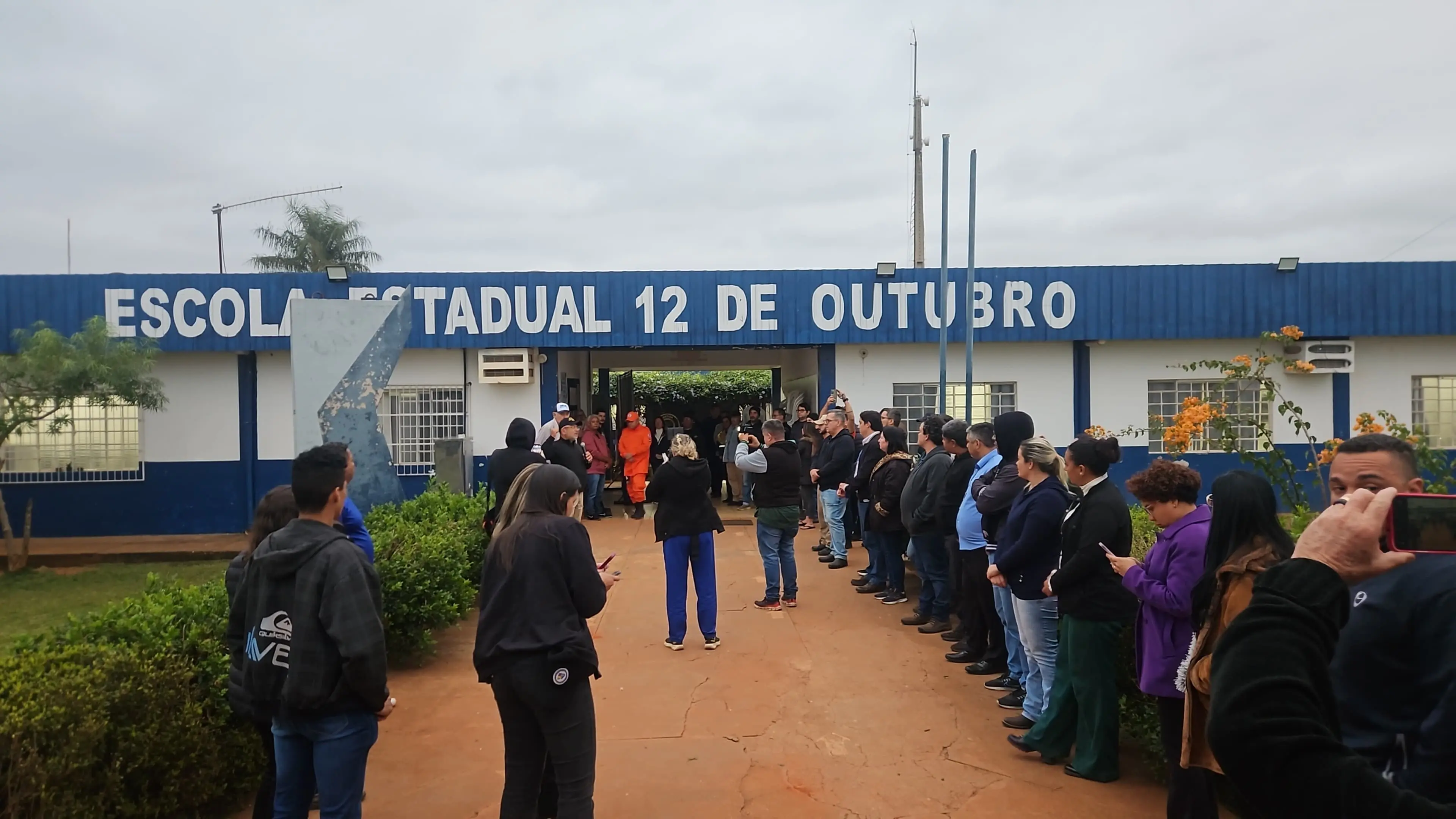 Pessoas se organizam em duas filas paralelas, formando corredor humano na entrada da Escola Estadual 12 de Outubro. Fachada azul e branca ao fundo, com flores e arbustos em destaque no jardim.