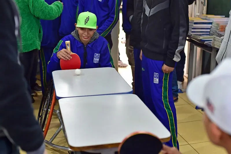 Jovem em cadeira de rodas joga tênis de mesa adaptado com outros colegas. Ele sorri enquanto rebate a bola com a raquete sobre duas carteiras escolares improvisadas como mesa