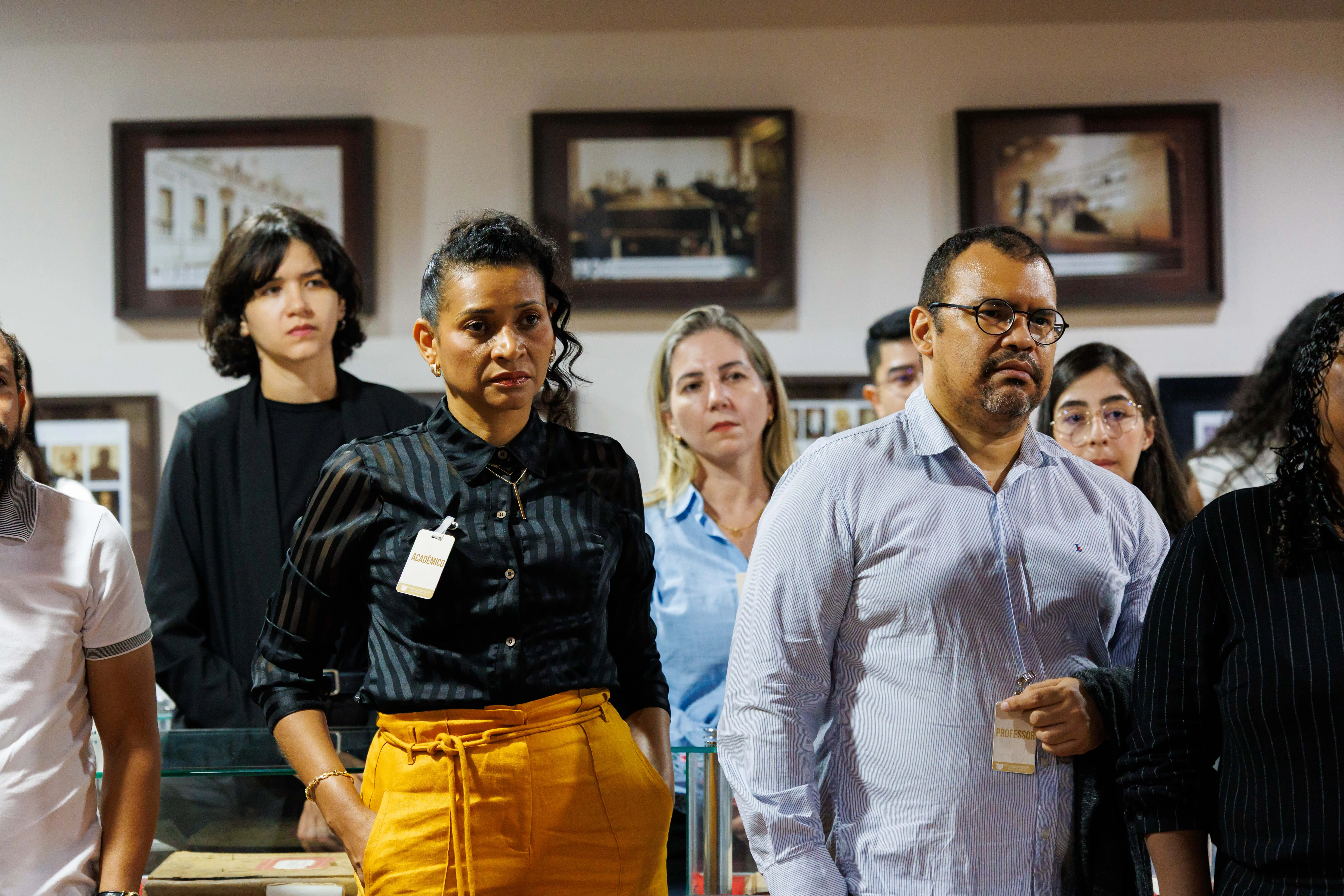 Grupo de pessoas observa atentamente uma atividade em espaço cultural. Em destaque, mulher com crachá e calça amarela e homem de óculos com crachá de professor. Ao fundo, quadros fotográficos emoldurados.