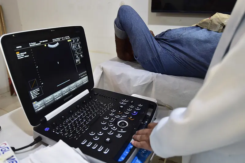 Foto que mostra a mão de um médico manuseando um monitor de ultrassom e, em segundo plano, o paciente deitado na maca com a mão sobre o rosto. Ele usa camisa jeans e o médico jaleco branco.