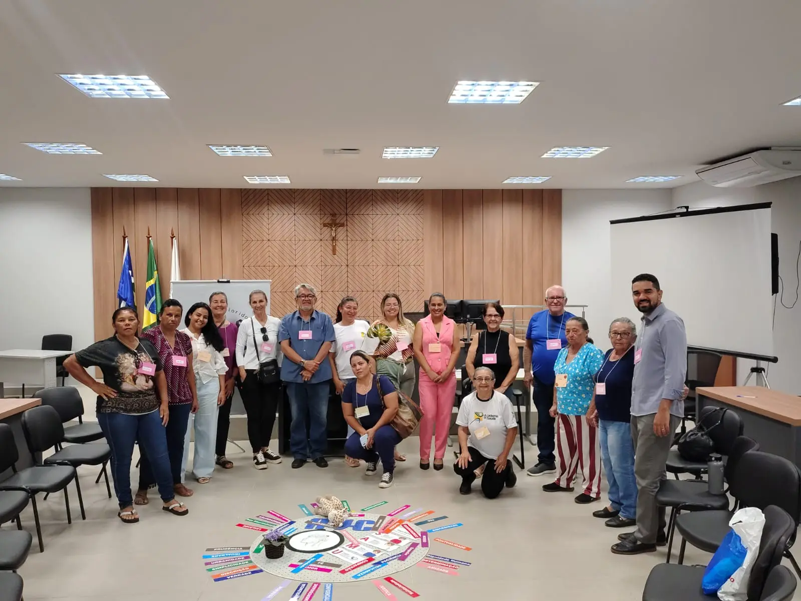 Grupo posando para foto em pé, sorridentes, ao redor do centro do Círculo de Paz. Ao fundo, bandeiras, painel de madeira e a estrutura de um plenário com crucifixo na parede.