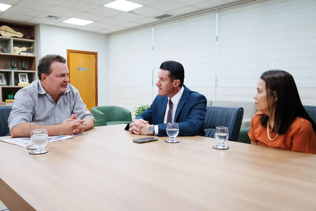 Em uma sala de reunião, o presidente da Assembleia Legislativa, Max Russi, o desembargador Rodrigo Curvo e a diretora da Ouvidoria do TJMT, Larissa Shymoia, estão sentados em volta de uma mesa, conversando.