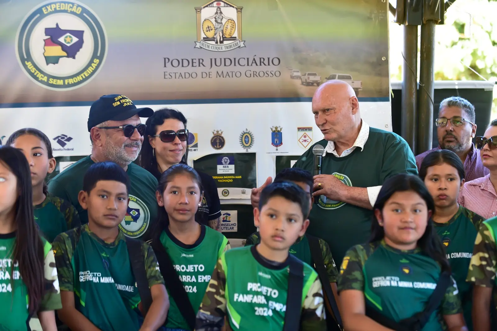 Autoridades e crianças reunidas na abertura da Expedição Justiça Sem Fronteiras. Ao fundo, banner com logotipos institucionais. Crianças usam camisetas verdes com inscrições de projetos sociais.