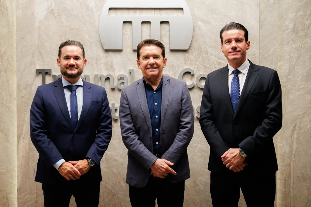 Da esquerda para a direita: juiz Bruno D'Oliveira, conselheiro de contas Sérgio Ricardo e desembargador Rodrigo Curvo posam sorrindo para a foto. Eles estão em pé e, atrás deles, está uma parede com a logomarca do TCE-MT em mármore.