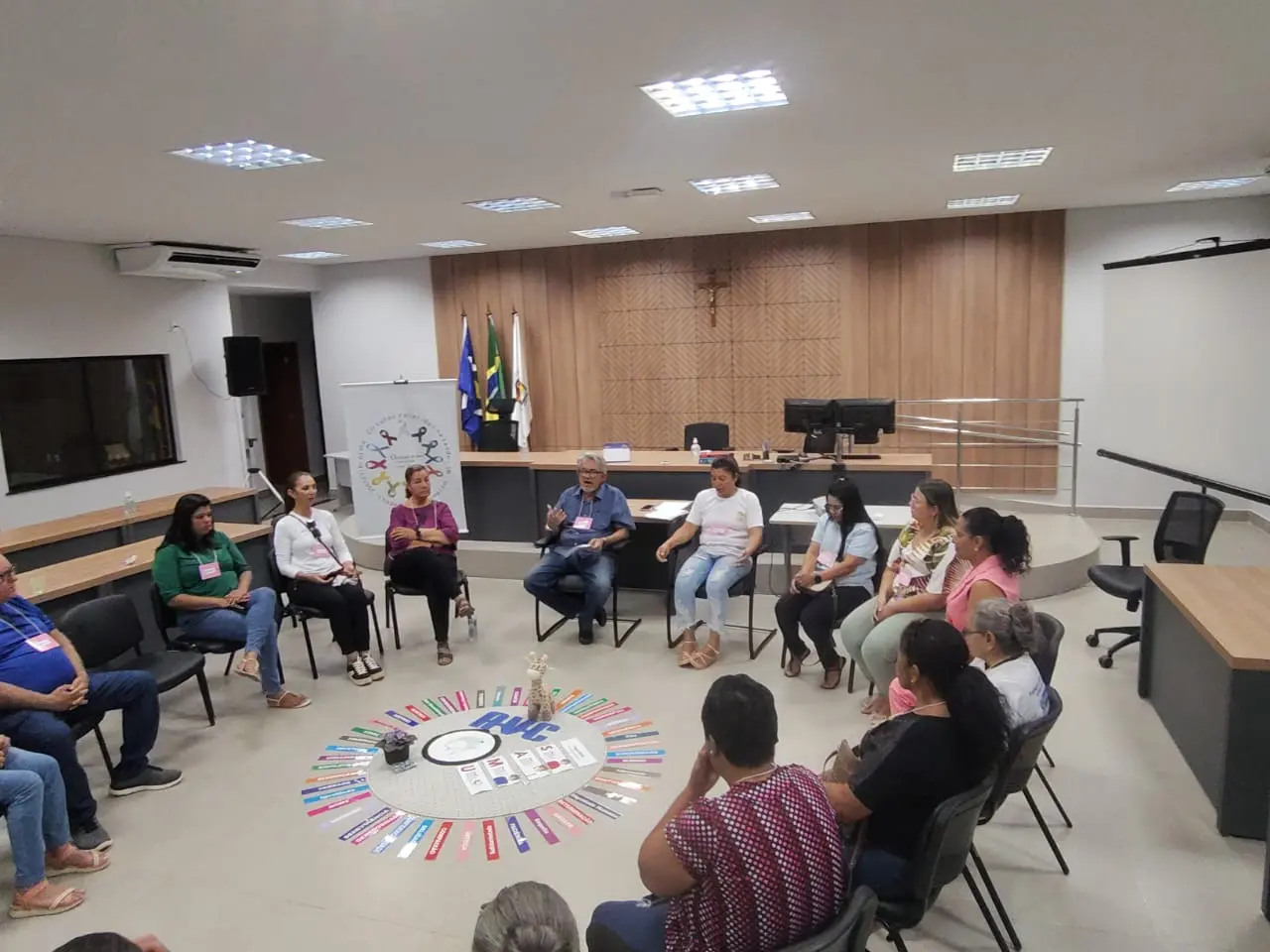 Círculo de diálogo com participantes sentados em roda, trocando experiências. Ao fundo, painel institucional com logomarca e bandeiras; clima de escuta e acolhimento é evidente.