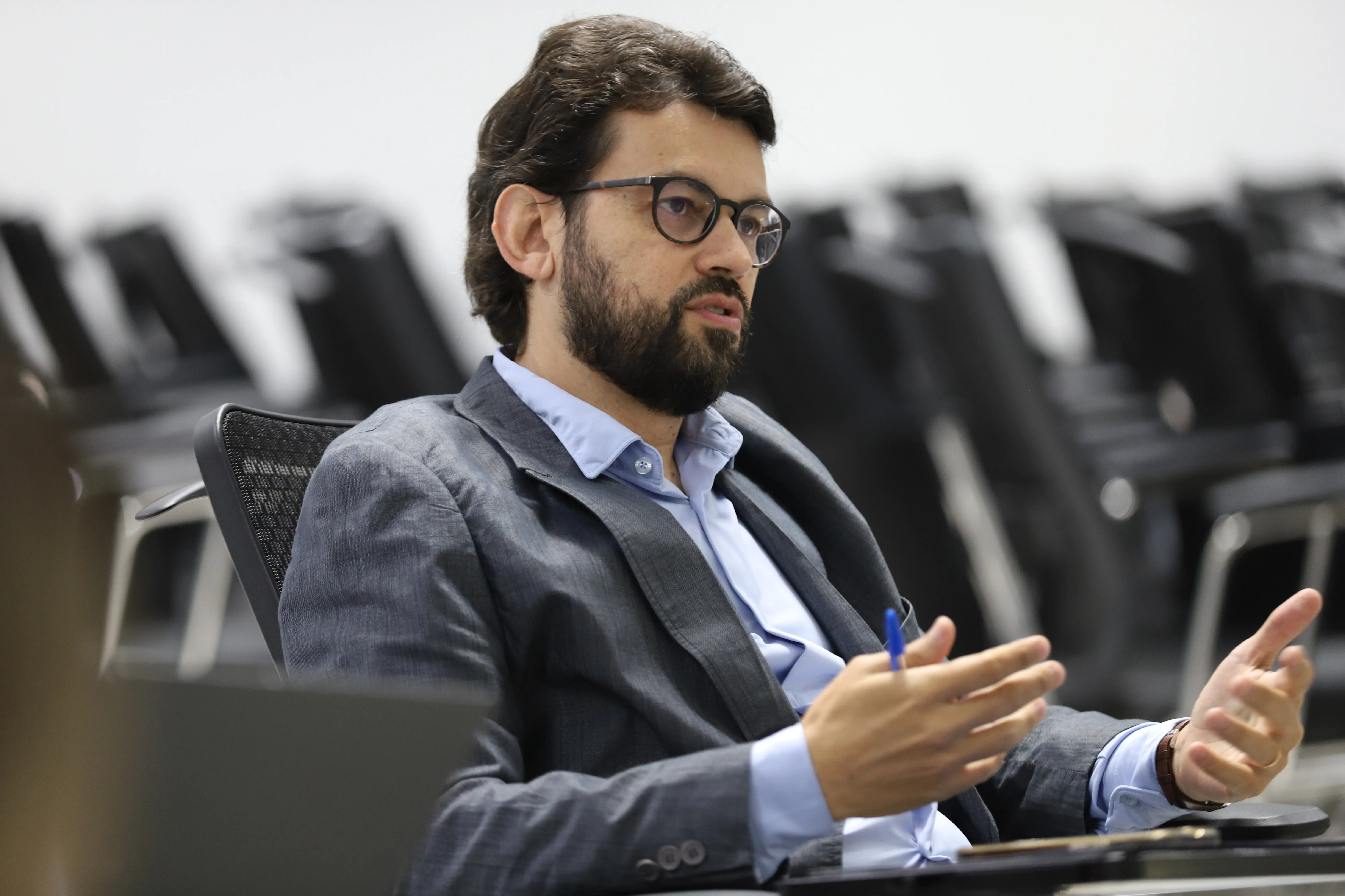 Um homem com barba, de óculos e terno, Tiago Abreu, está sentado, gesticulando enquanto fala. Ele parece estar em uma reunião ou apresentação, com cadeiras desfocadas ao fundo.