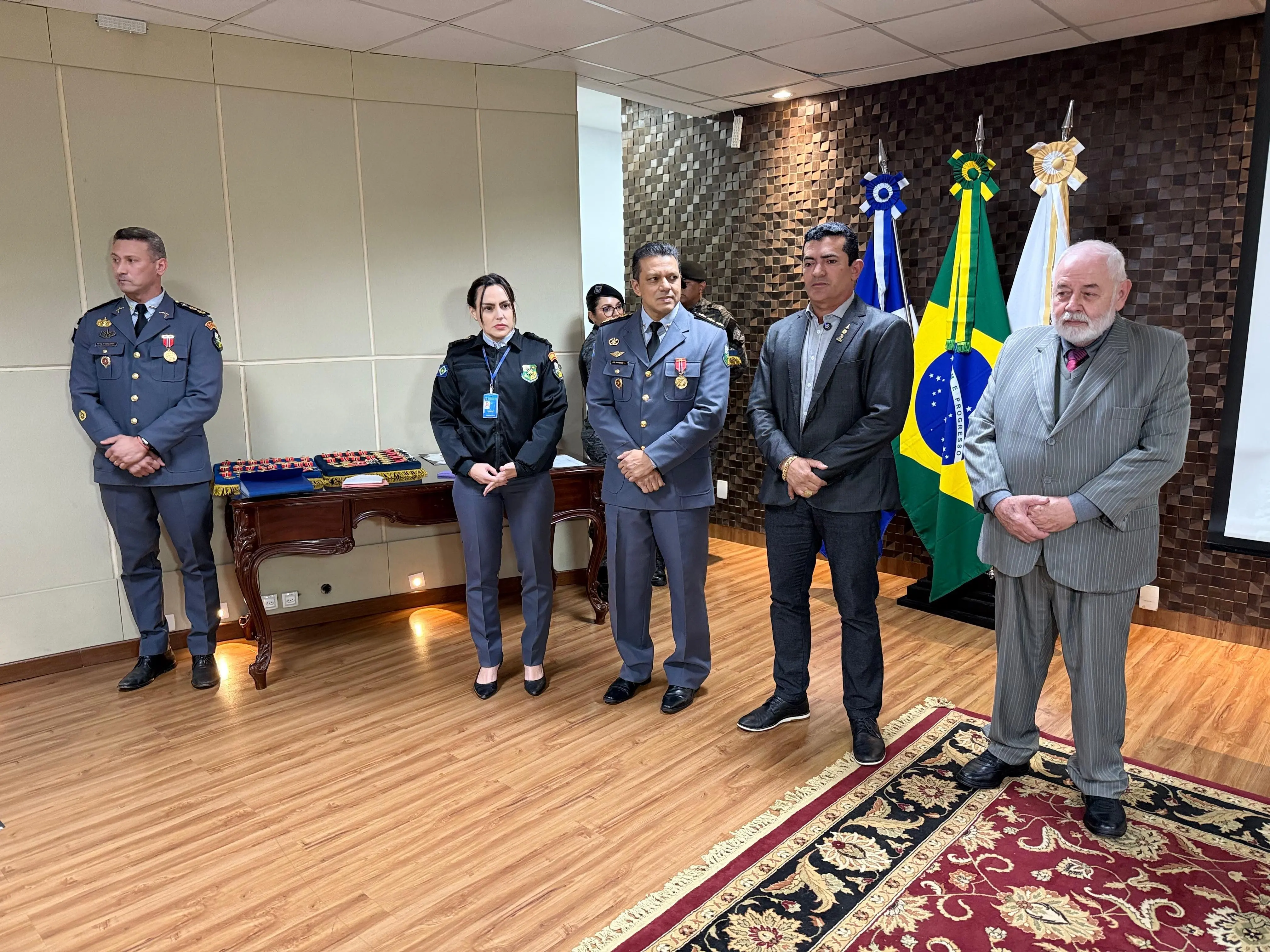 Seis pessoas em pé em cerimônia oficial. Entre elas, militares fardados, uma mulher com uniforme e um homem idoso de terno cinza. Ao fundo, bandeiras do Brasil, de Mato Grosso e institucional.