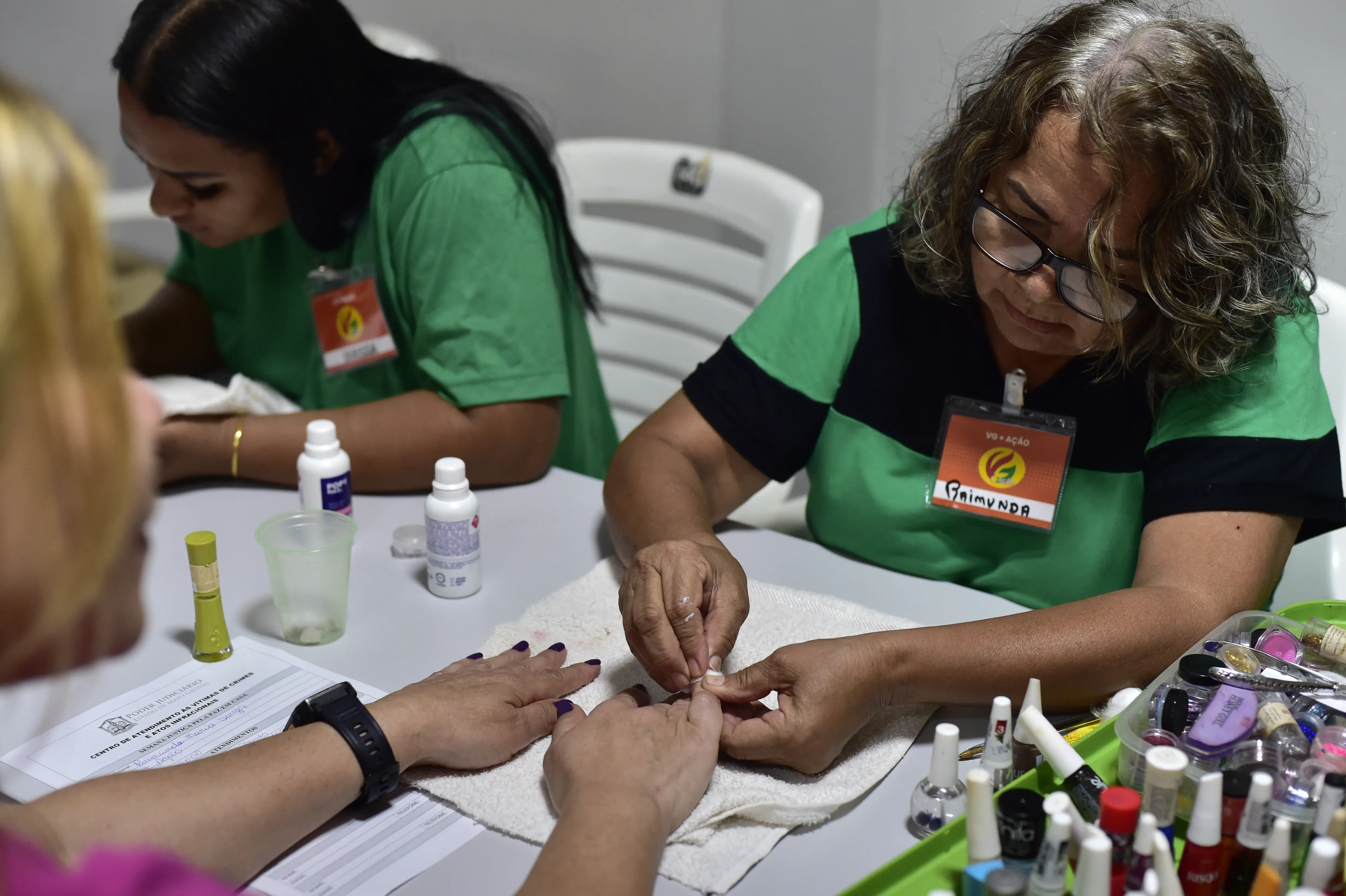a imagem mostra uma manicure, que veste camiseta verde com detalhes em preto, fazendo a unha de uma mulher. Somente as mãos da mulher aparecem. 