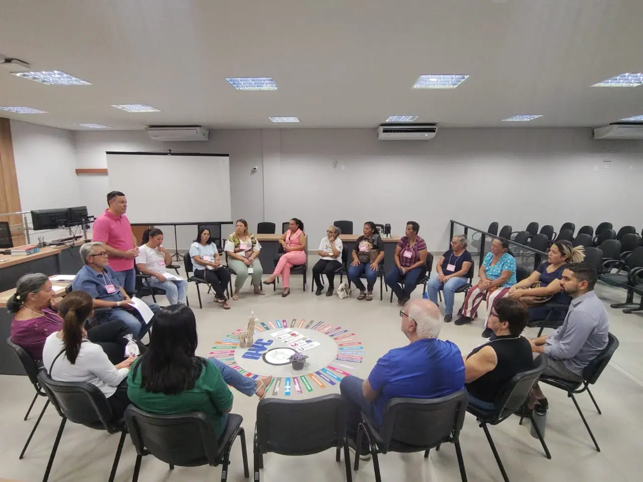 Participantes sentados em círculo, atentos à fala de um dos presentes. O ambiente é uma sala ampla, com ar-condicionado, projetor, cadeiras e estrutura de plenário ao fundo.
