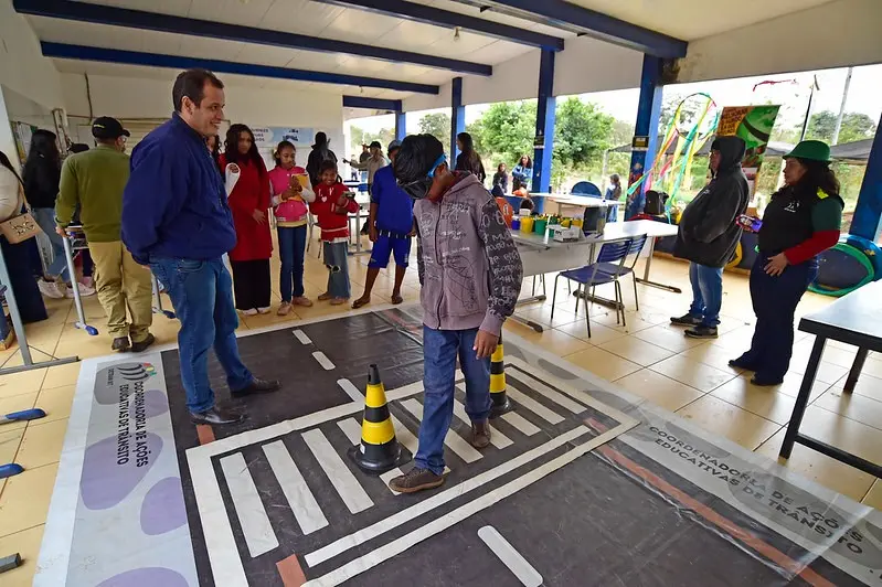 Criança atravessa simulação de faixa de pedestres usando óculos que distorcem a visão. A atividade educativa é acompanhada por adultos e promove conscientização sobre segurança no trânsito de forma lúdica.