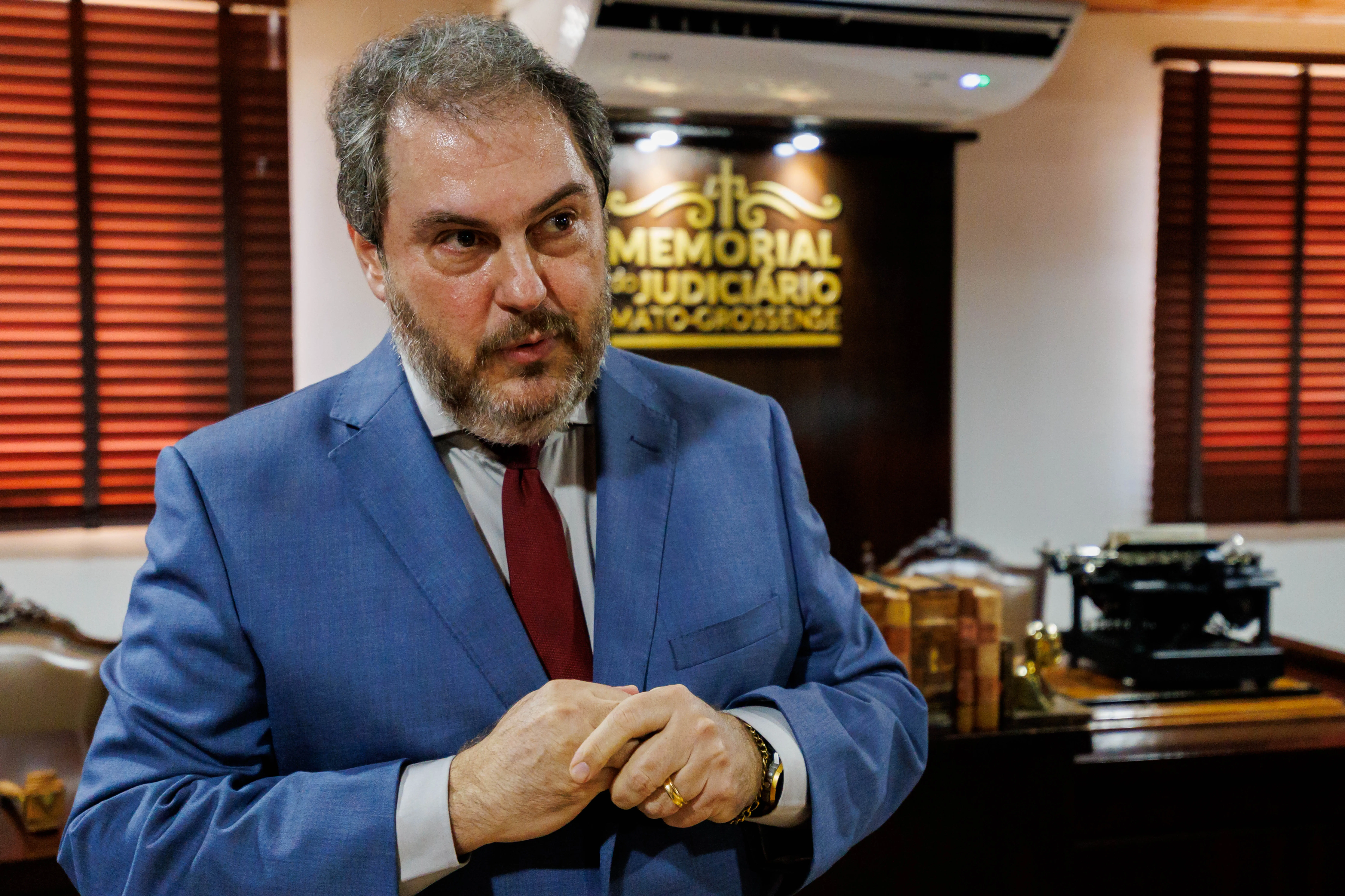 Desembargador Wesley Sanchez usa terno azul e gravata vermelha fala em ambiente solene do Memorial do Judiciário Mato-grossense. Ao fundo, livros antigos, máquina de escrever e placa institucional reforçam o caráter histórico.