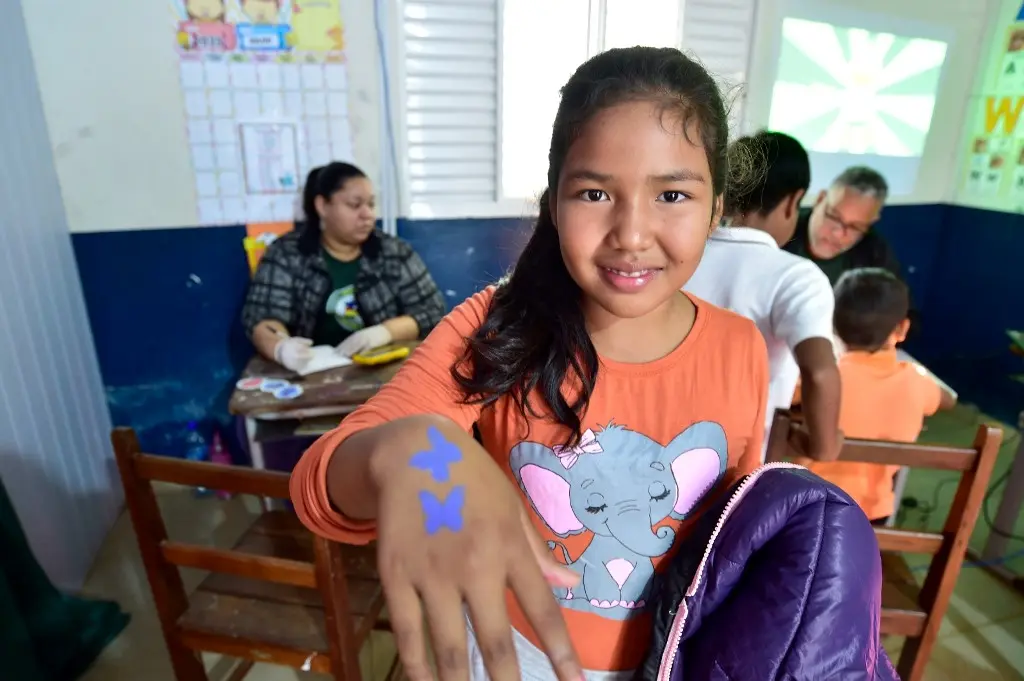 Menina sorrindo exibe a mão com a pintura de duas borboletas lilás. Ela está em uma sala de aula. Atrás dela, há mais crianças sendo atendidas pelos servidores da Secel.