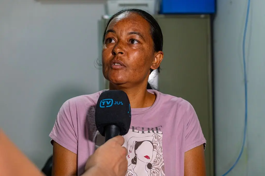 A dona de casa Elaine Cristina Bispo de Jesus concede entrevista à TV.Jus. Ela é uma mulher negra, de cabelos lisos, presos e pretos, olhos escuros, usando camiseta lilás estampada.