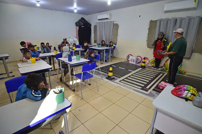 Sala de aula onde ocorre atividade pedagógica do Detran. No chão, há um tapete representando o trânsito. Alunos estão nas carteiras observando.
