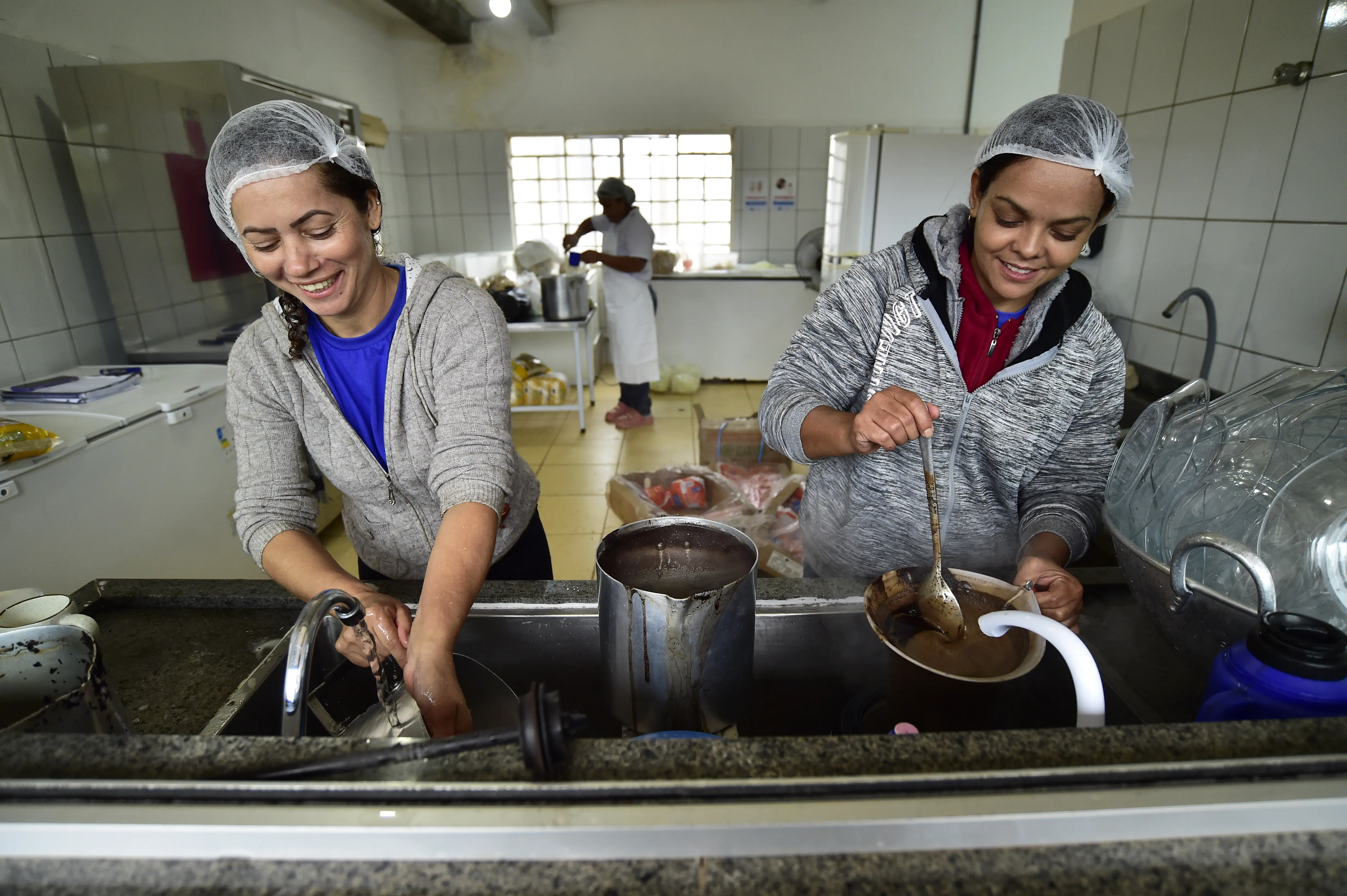 Foto que mostra, em primeiro plano, duas mulheres lavando louça e preparando café e, em segundo plano, outar preparando comida, na cozinha da escola onde ocorreu o mutirão da Justiça Sem Fronteiras. Elas sorriem enquanto trabalham.