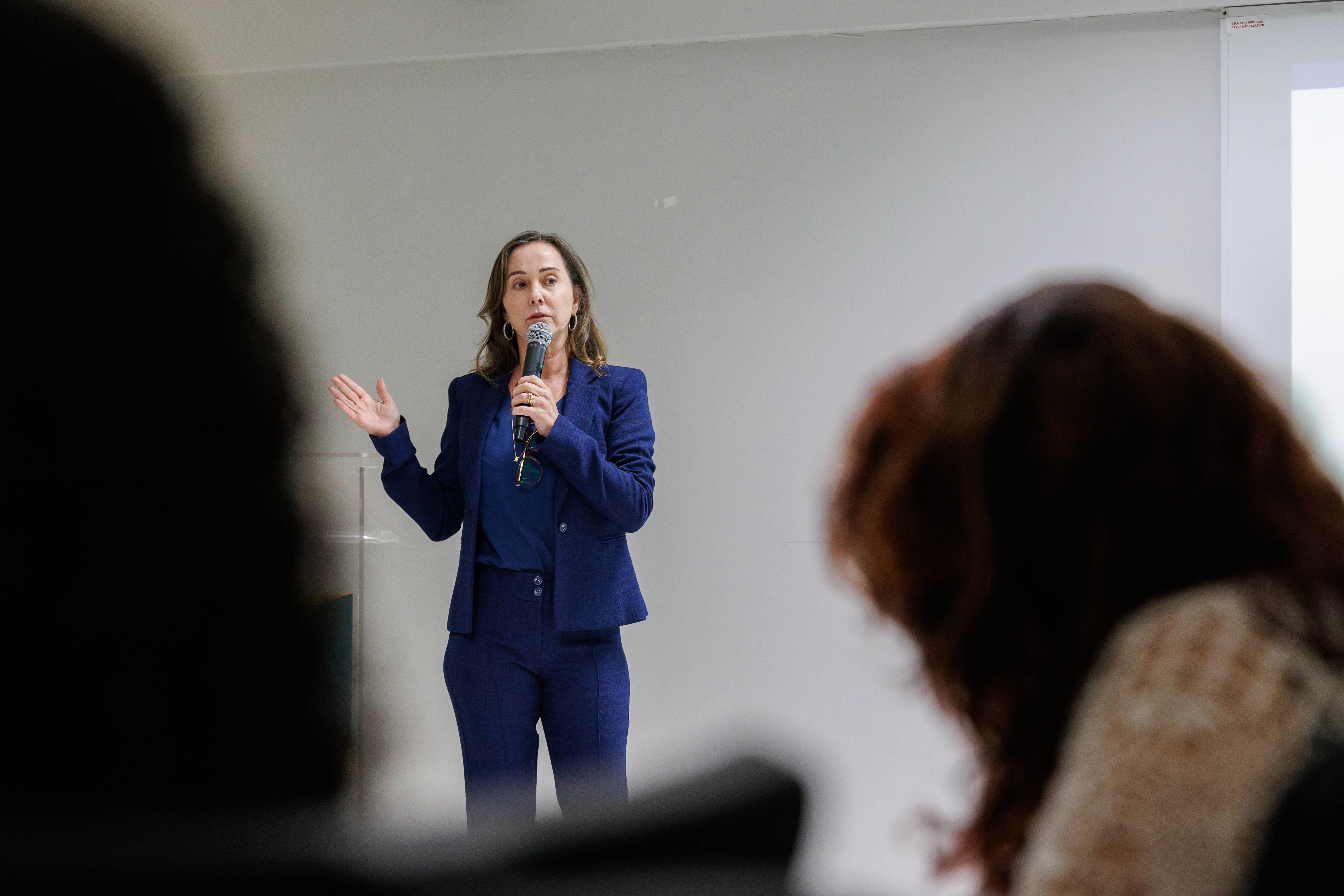 Juíza Leilamar de terno azul fala ao microfone em uma sala clara, gesticulando com a mão esquerda. À frente dela, pessoas acompanham atentamente, com parte de suas silhuetas desfocadas em primeiro plano.