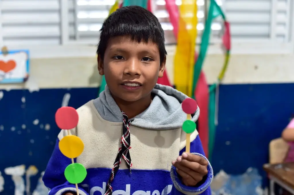 Menino negro, de olhos pretos e cabelos lisos e pretos sorri para a foto, mostrando dois semáforos que fez com palito e papeis coloridos.
