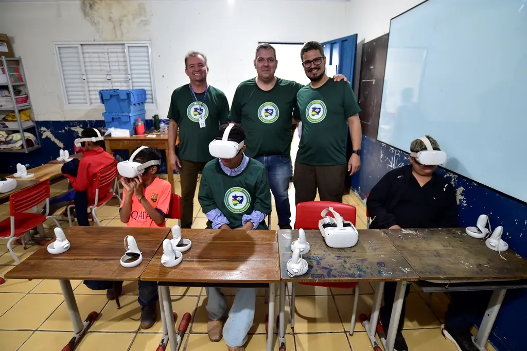 Sala de aula com três crianças utilizando óculos de realidade virtual. Três servidores da Secretaria de Ciência e Tecnologia posam para a foto sorrindo.