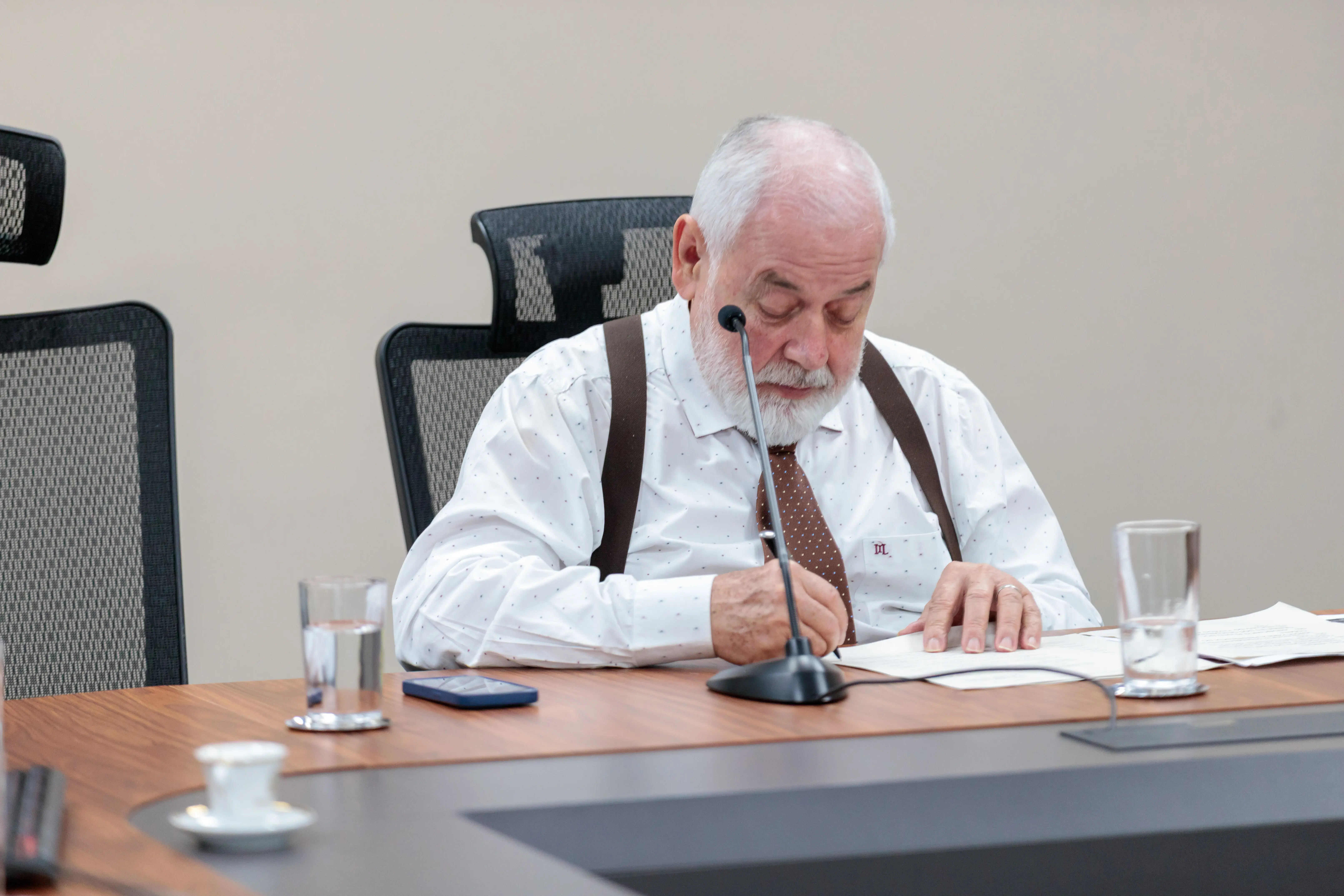 Presidente Zuquim está sentado assinando documento. Ele é um homem branco, com barba e cabelos grisalhos. Usa camisa de manga comprida branca, suspensório e gravata marrons