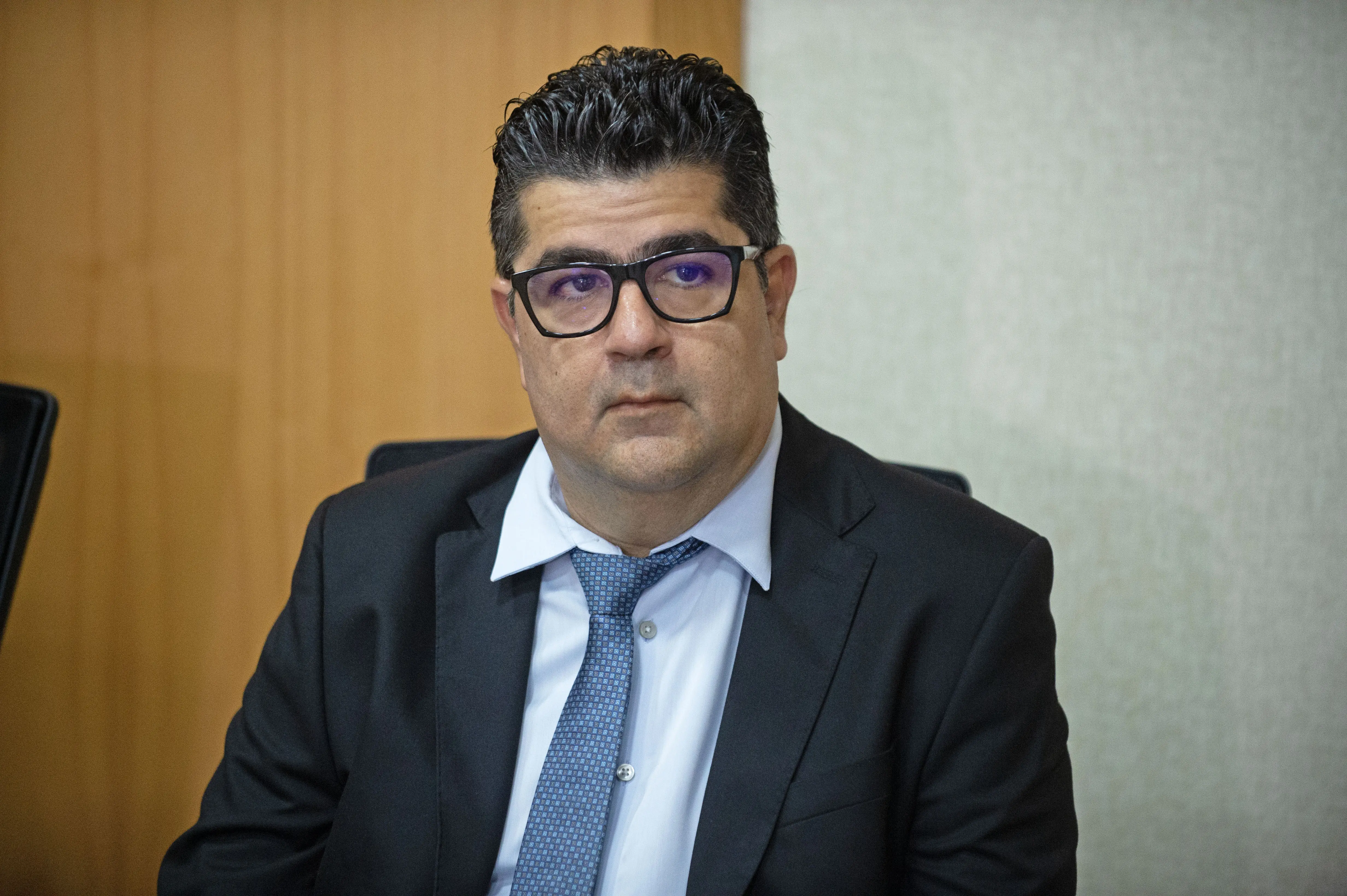 Juiz Marcos Terêncio sentado. Ele é um homem branco, de cabelos e olhos escuros, usando camisa azul clara, gravata azul, terno preto e óculos de grau de armação preta.