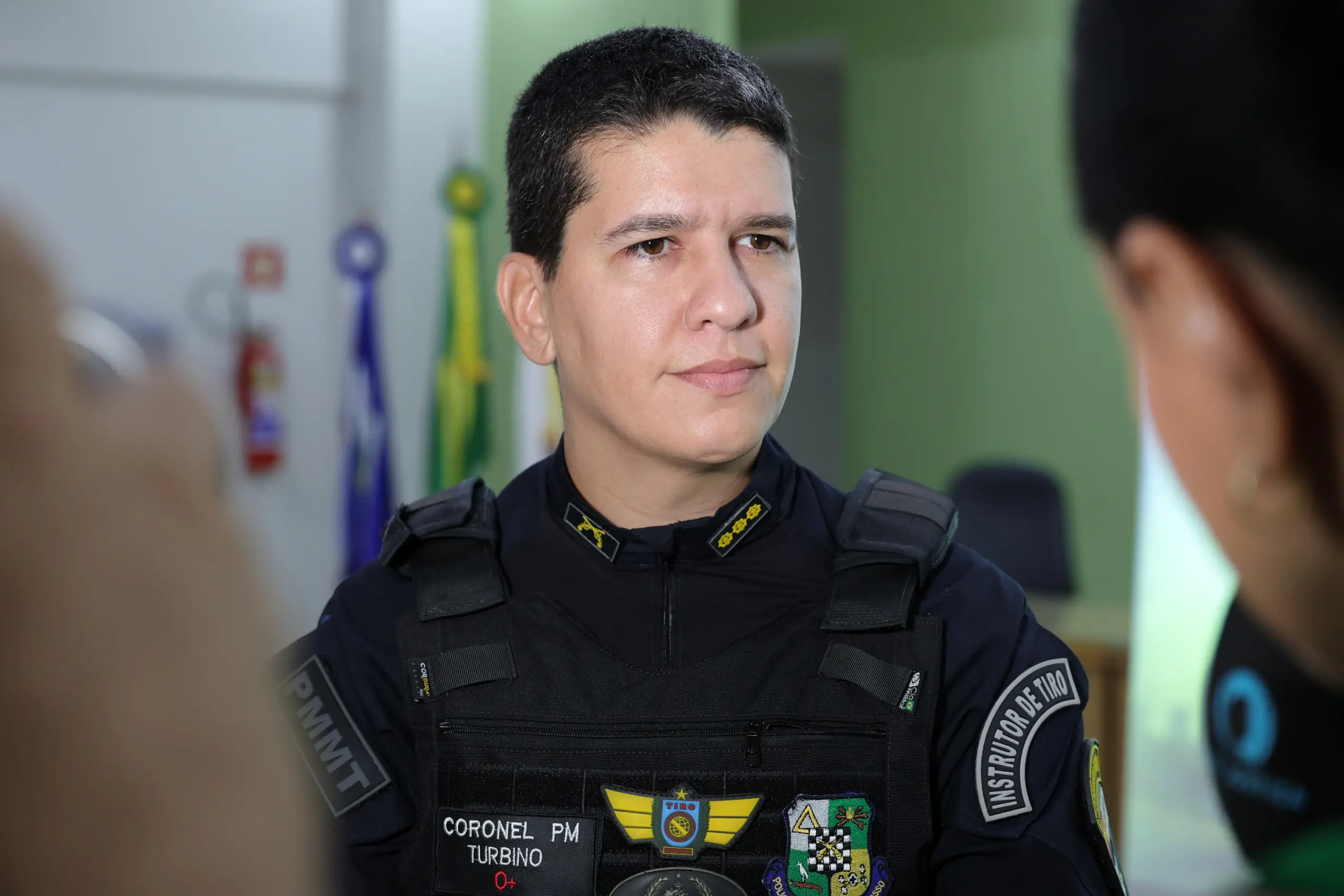 Primeiro plano de um homem fardado da Polícia Militar de Mato Grosso (PMMT). No colete há a inscrição "Coronel PM Turbino" e "Instrutor de Tiro". Ele olha para a direita de forma séria.