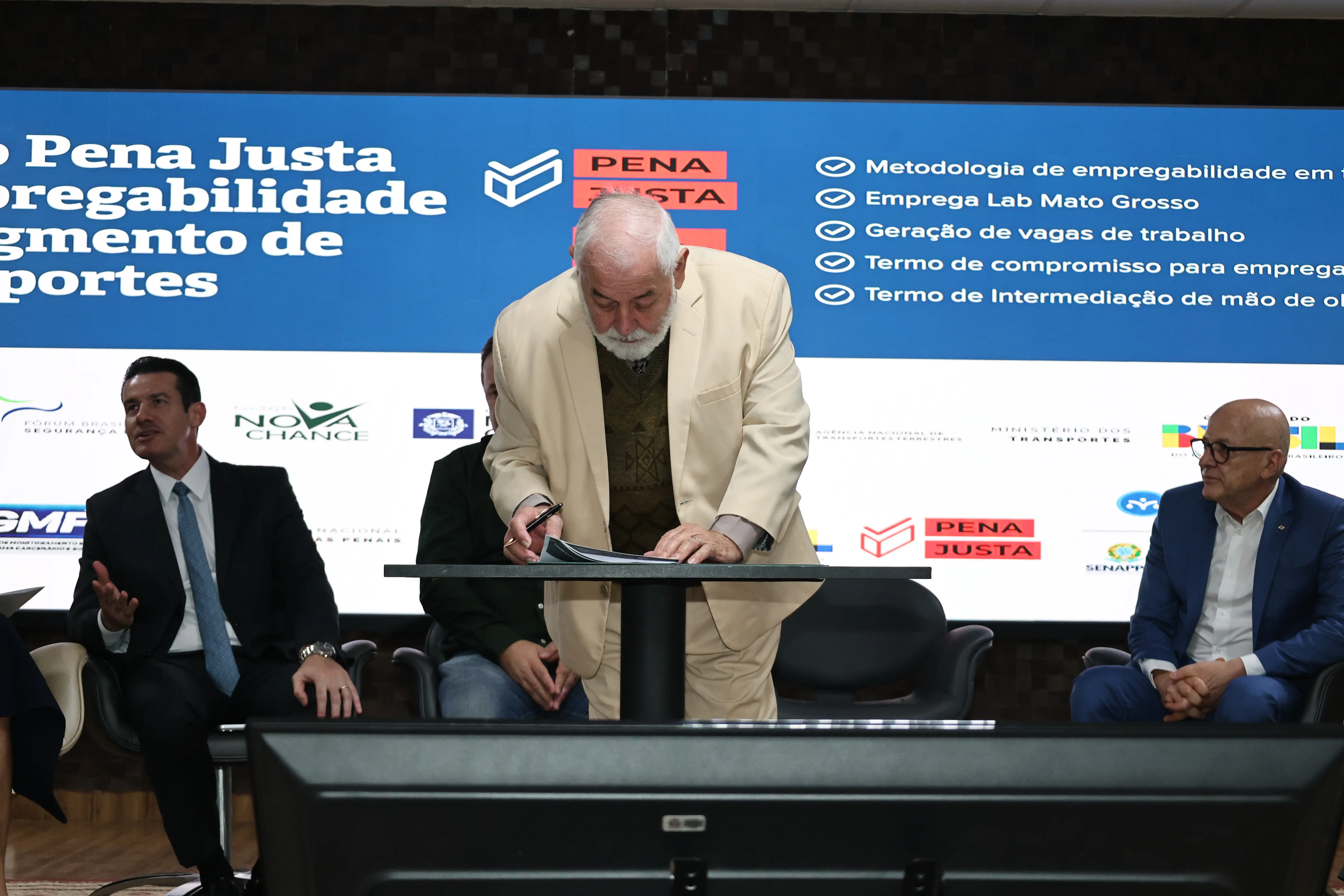 Homem branco, de terno bege inclinado sobre uma mesa, com uma caneta para assinatura de documentos. Ao fundo, telão exibe o texto "Pena Justa Empregabilidade", outros três homens aparecem na cena.