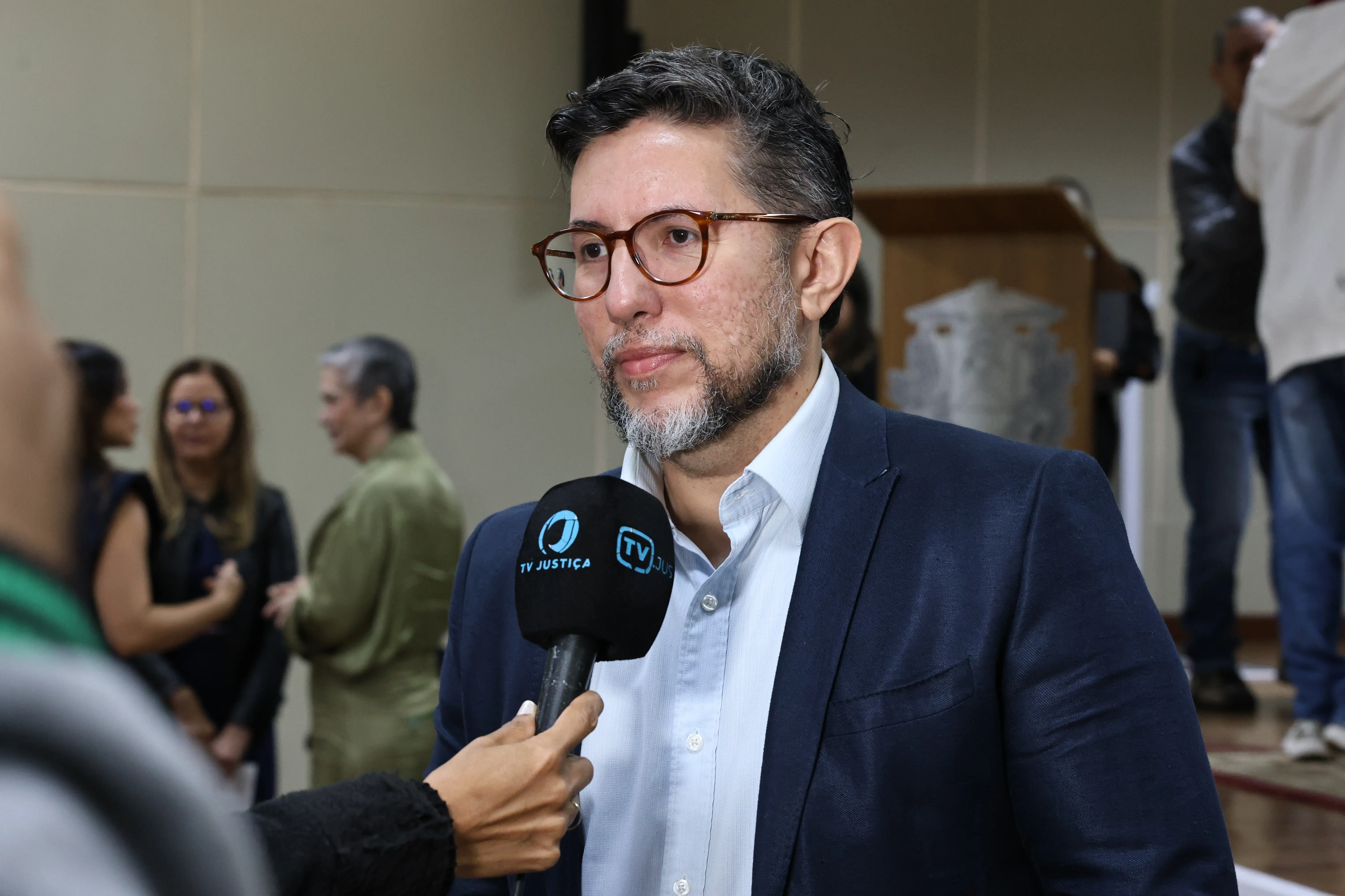 Homem branco, com cabelo preto e barba grisalha. Usa terno azul marinho, com camisa azul claro. Ele fala a um microfone, que tem escrito em azul "TV Justiça" e "TV Jus"