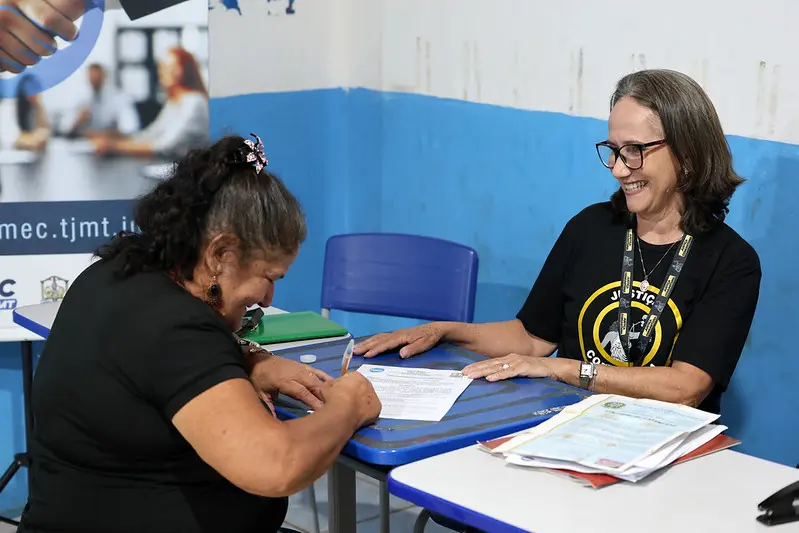 Foto horizontal colorida, em plano médio, que mostra Sônia Maria Castelari sorrindo, enquanto assina termo de audiência de divórcio. Sentada de frente para ela, está a mediadora do Cejusc de Paranatinga, Andreia Correa, também sorrindo. Ao fundo, há um banner do Cejusc. 