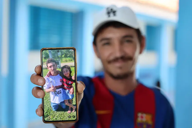 Foto horizontal colorida, em plano fechado, que mostra Wellington Hartman, desfocado, mostrando a tela do celular (em primeiro plano), onde aparece a foto dele com a filha, Marissol. Ele sorri.