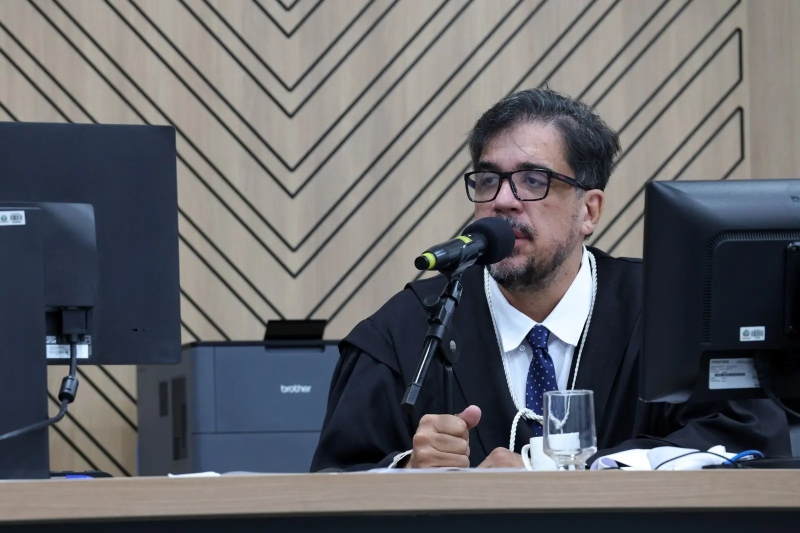Foto horizontal colorida que mostra o juiz Marcos Faleiros sentado em frente a um computador, durante sessão do Tribunal do Júri, no Fórum de Cuiabá. ele é um homem branco de cabelos preto, usando toga e óculos de grau.