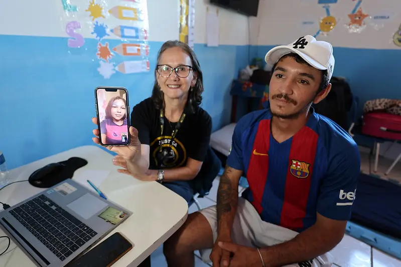 Foto horizontal colorida, que mostra a mediadora do Cejusc de Paranatinga, Andreia Correa, e o metalúrgico Wellington Hartman sentados em frente a um notebook, durante audiência de reconhecimento de paternidade. Andreia mostra a tela do celular, onde a mãe da criança reconhecida participa da sessão.