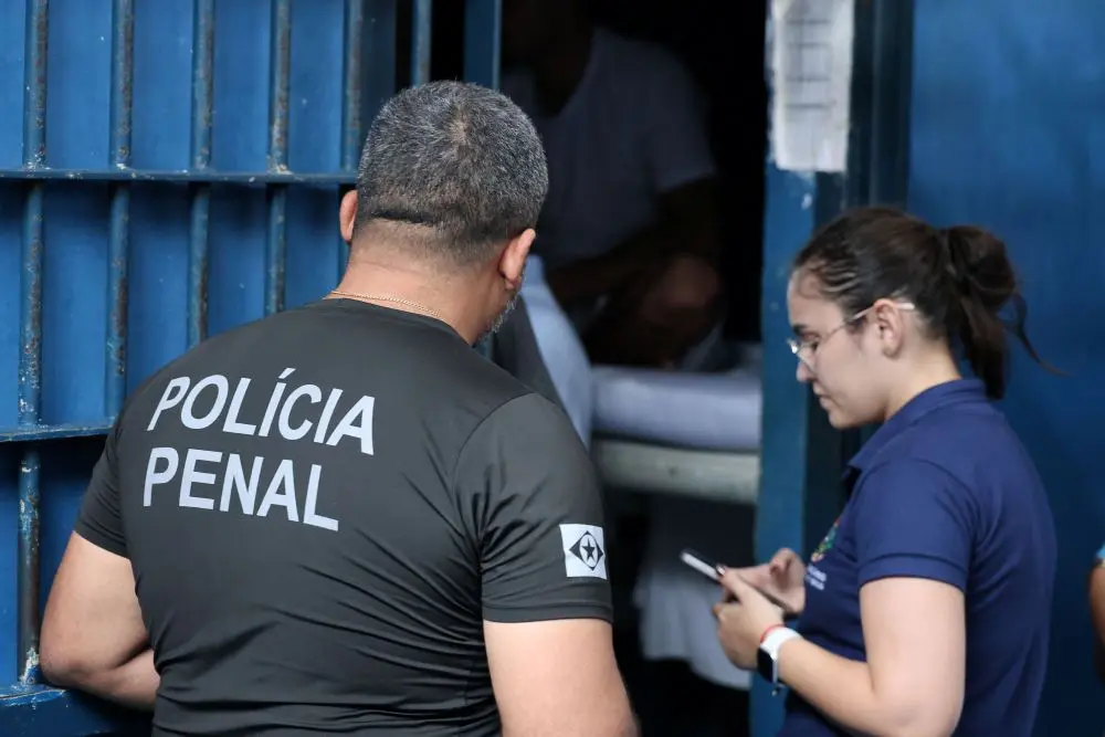 Foto horizontal colorida, em plano médio, que mostra um policial penal de costas, na porta de uma cela, ao lado de uma servidora do Estado.