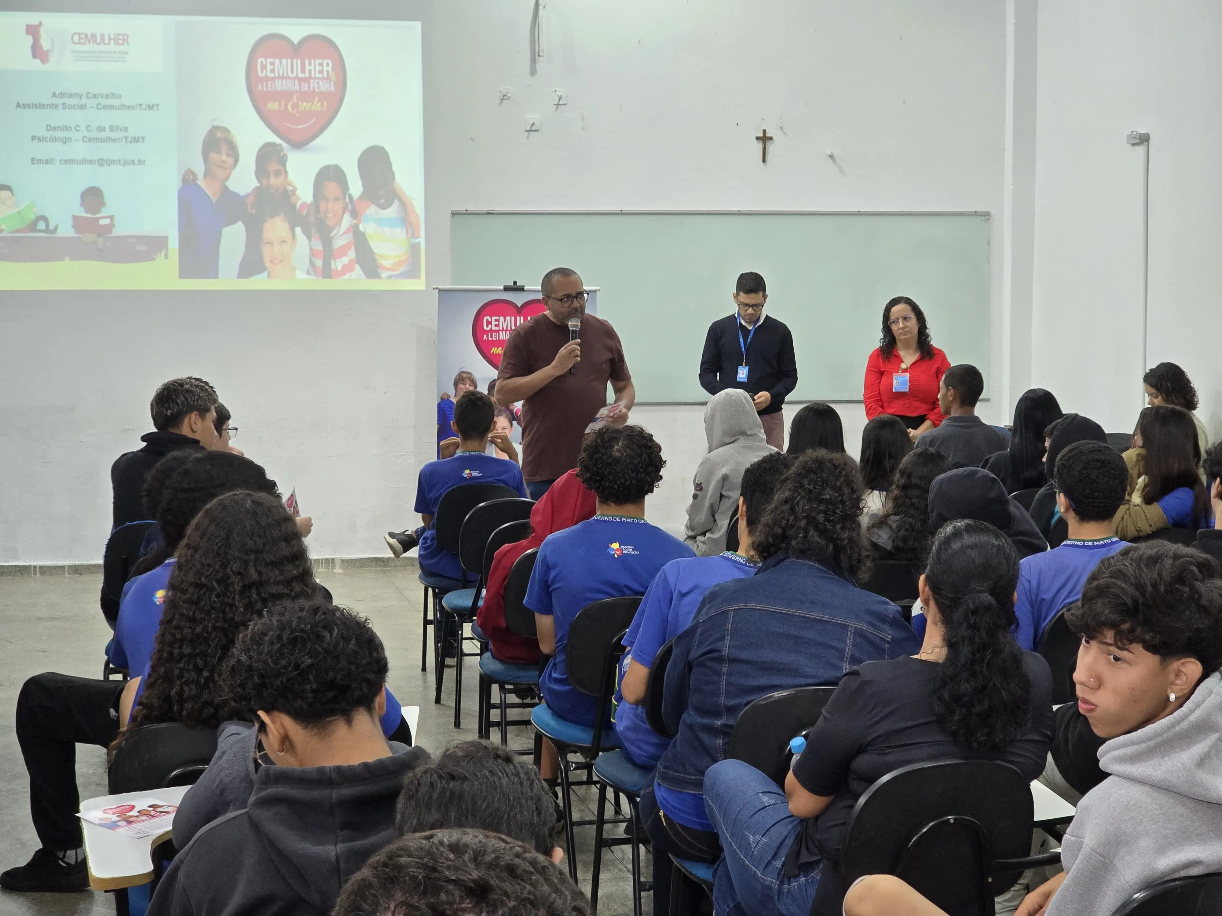 A imagem mostyra primeiro plano de vários alunos sentados em cadeiras, dentro da sala de aula, usando uniformes azuis. De frente apra eles, em pé, estão o diretor da Escola, psicólogo e a assistente social da Cemulher