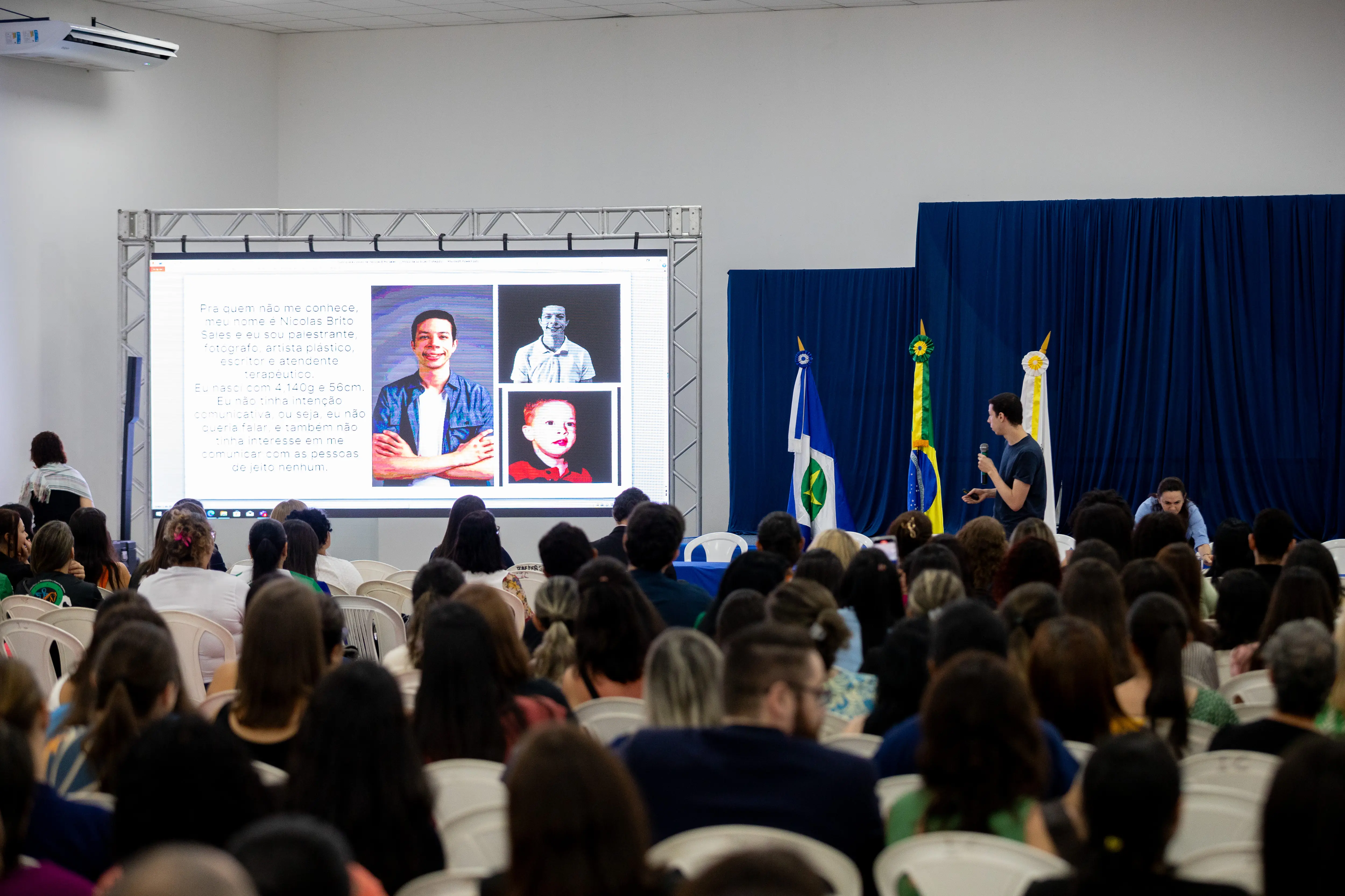 imagem em ân gulo aberto de pessoas sentadas assistindo palestra durante o MT Inclusivo em Sorriso. ao fundo, no palco, está Nicolas e ao lado um telão onde passa slide
