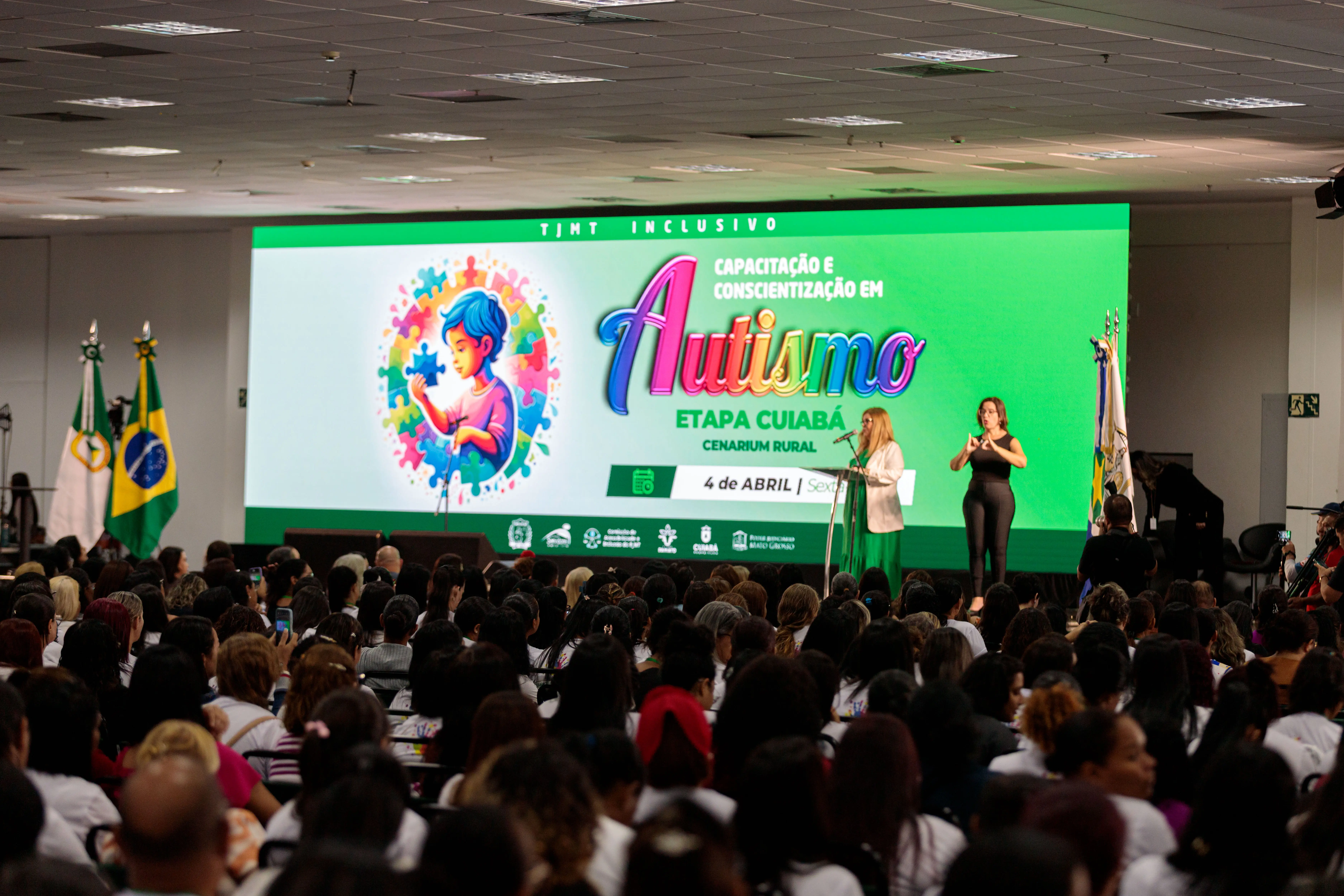 04-	Imagem da desembargadora Nilza Maria, uma mulher parda, cabelos loiros e lisos. Ela veste uma blusa verde e sobre ela um terno branco. Ao fundo, um painel de LED com a arte do evento. Fundo verde, uma criança envolva em quebra-cabeças e a palavra autismo em destaque, com letras coloridas
