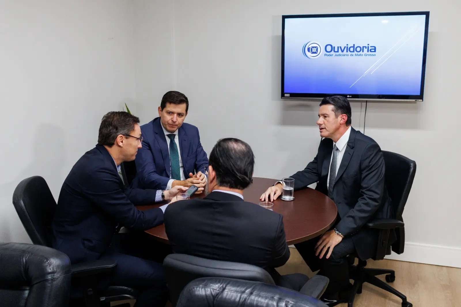 Imagem registra quatro homens envolta de uma mesa redonda de madeira. Eles estão em reunião e vestem roupas formais, ternos escuros e gravata. Ao fundo há um monitor que reproduz uma imagem com a escrita "Ouvidoria".