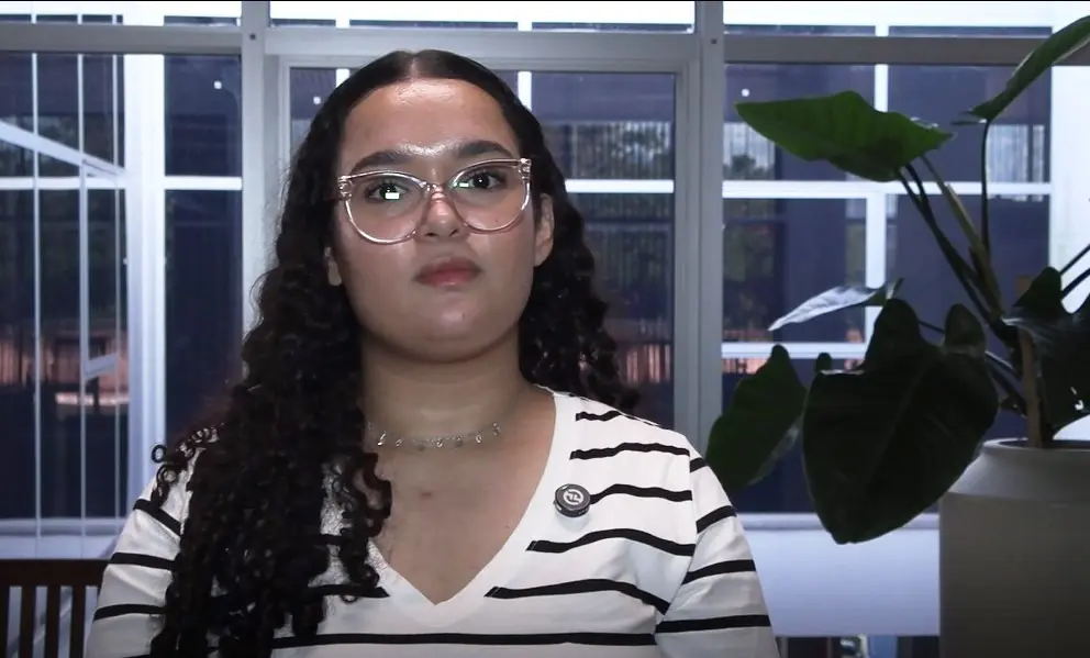 A imagem mostra a acadêmica Giovanna. Ela é uma jovem de pele clara, cabelos encaracolados, longos e escuros. Tem olhos escuros e usa óculos de grau. Esta vestindo uma camiseta branca com listras horizontais pretas