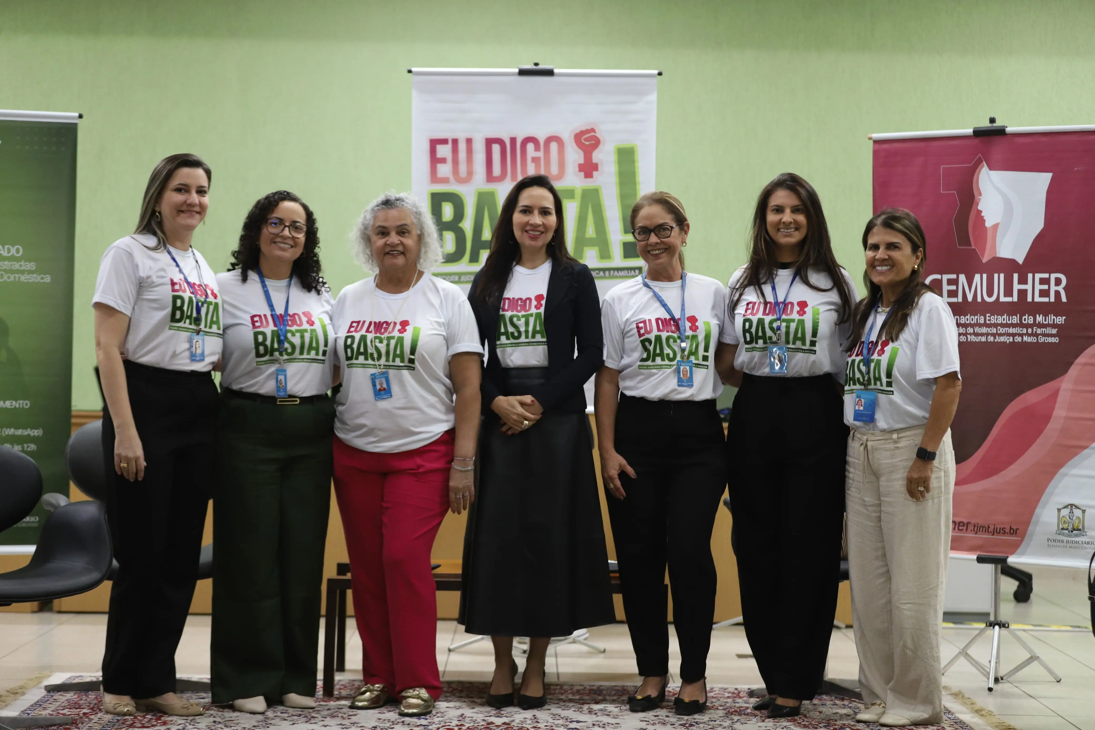 Grupo de oito mulheres posa sorrindo no palco. Todas vestem camisetas “Eu Digo Basta!” e crachás. Ao fundo, banners verdes e vermelhos destacam a campanha e a CEMULHER.