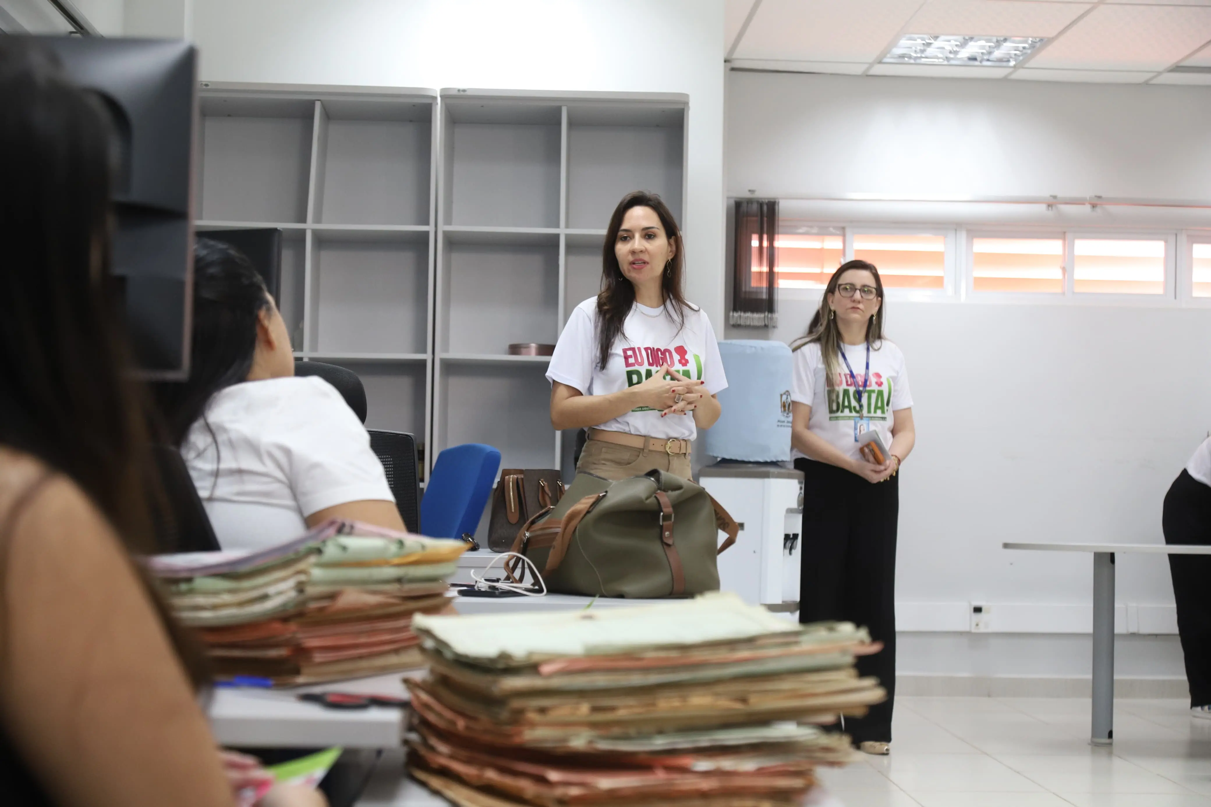Em um escritório em Jaciara, Mato Grosso, duas mulheres de camisetas "Eu Digo Basta!" estão de pé. Uma delas, no centro, fala para um grupo sentado, indicando uma apresentação da campanha. O local é iluminado, com estantes e mesas de trabalho ao fundo.