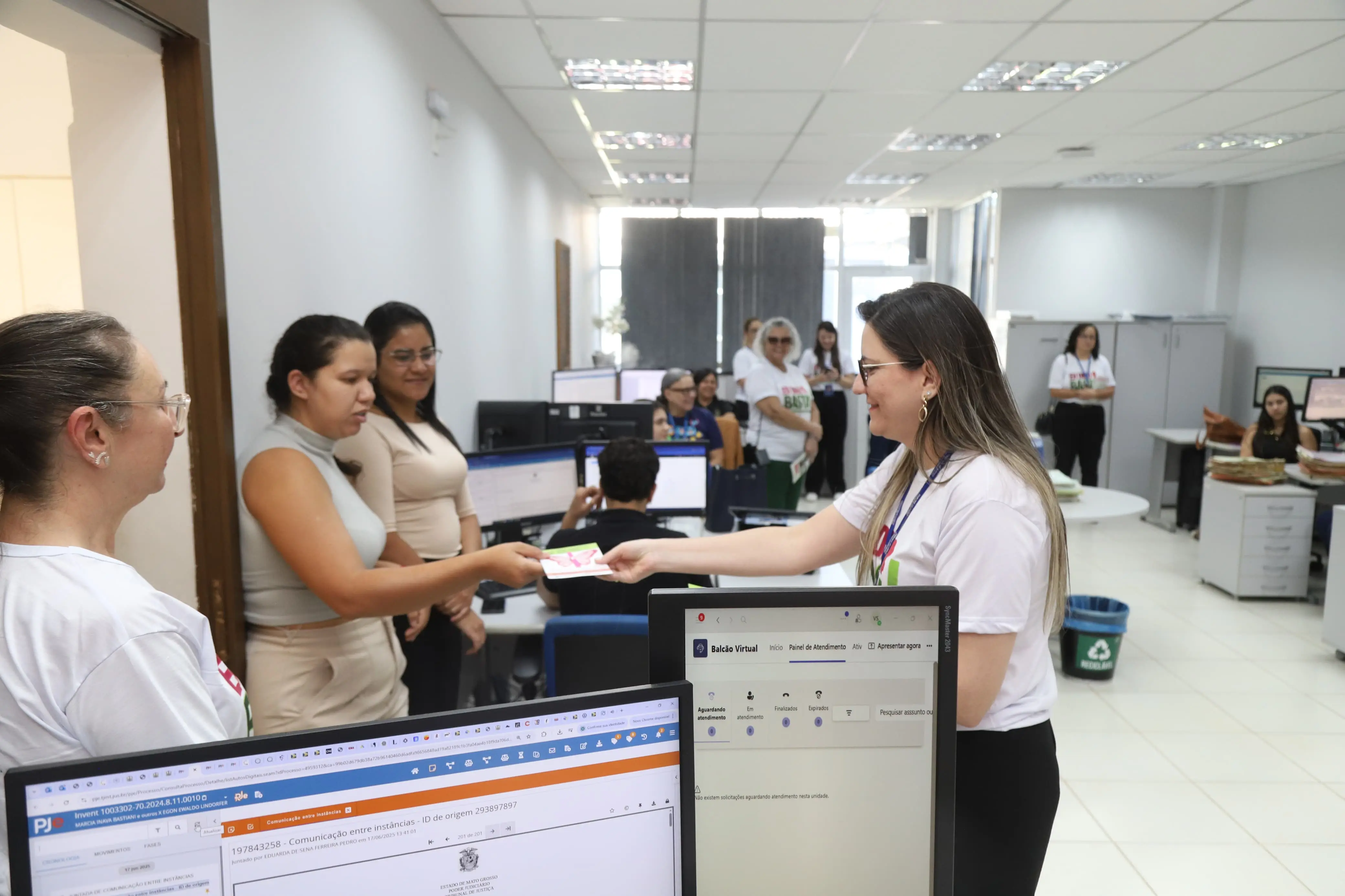 Jaciara, uma mulher de óculos e camiseta branca "Eu Digo Basta!" entrega um folheto a duas outras mulheres, que o recebem com atenção. O escritório é movimentado, com computadores e pessoas trabalhando ao fundo, ilustrando a distribuição de informações da campanha.