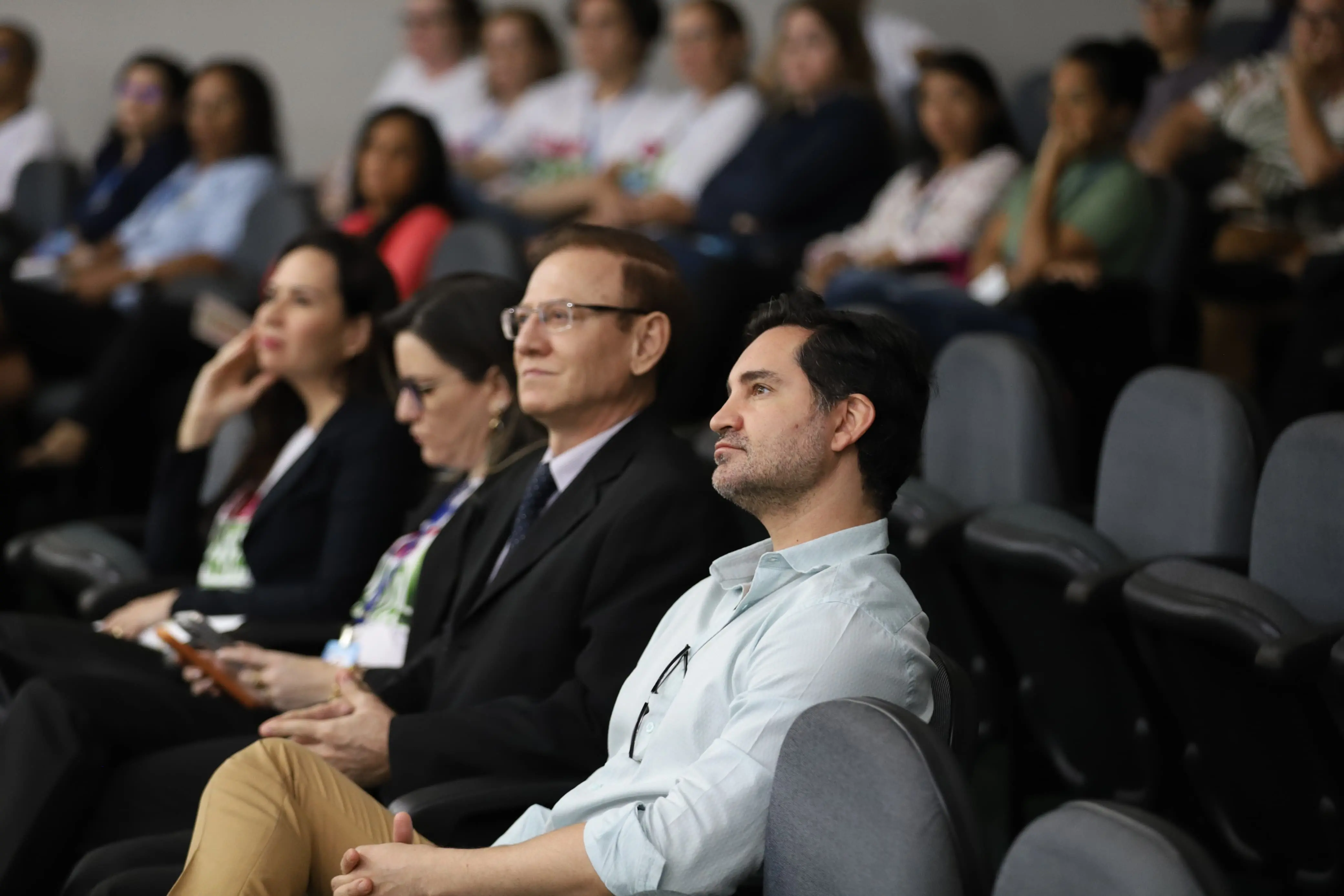 Homens e mulheres sentados no auditório assistem à palestra. Olhares atentos e concentrados voltados ao palco. Cadeiras cinzas preenchem o espaço ao fundo.