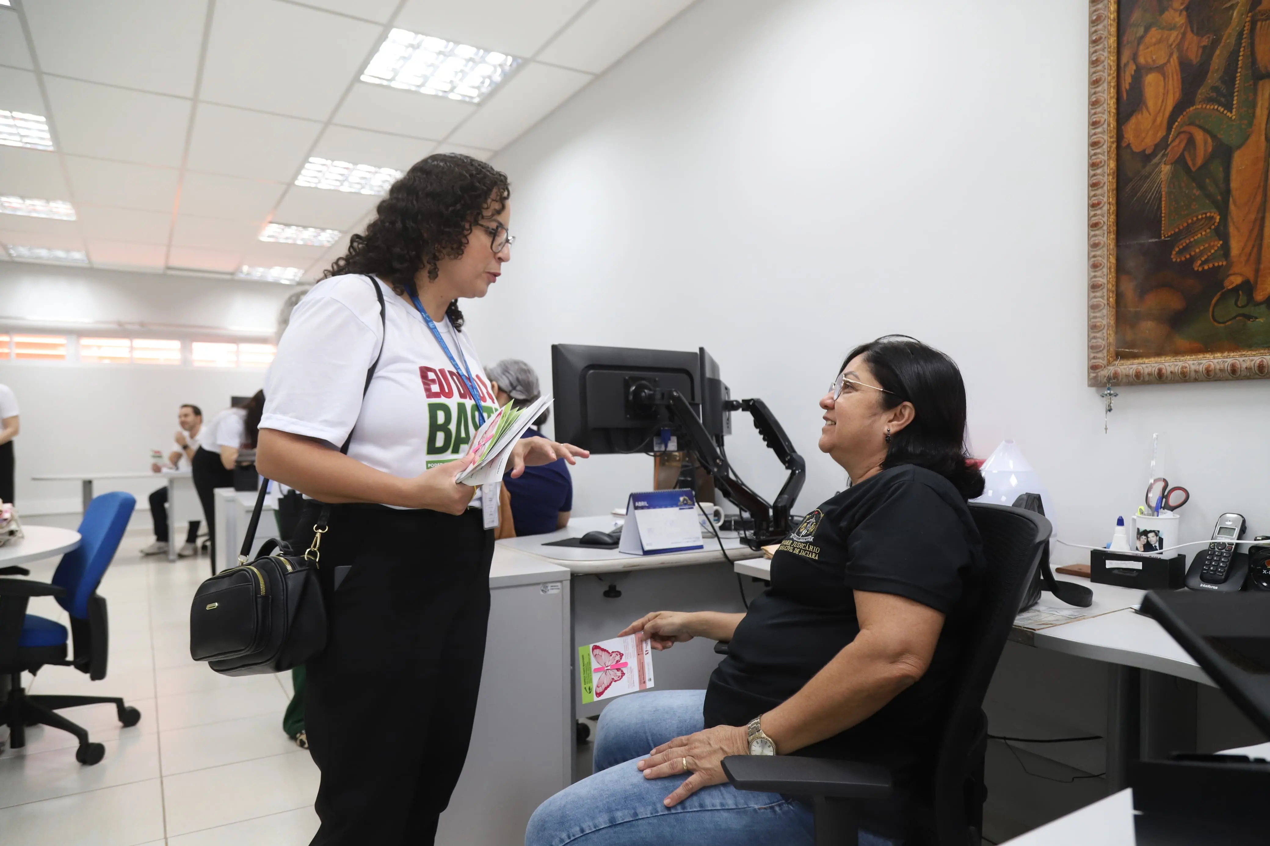 No interior do prédio em Jaciara, Mato Grosso, uma mulher de camiseta "Eu Digo Basta!" e cabelos cacheados escuros, em pé, dialoga com outra mulher sentada à mesa de um escritório. A mulher sentada, de óculos, veste uma camiseta escura 