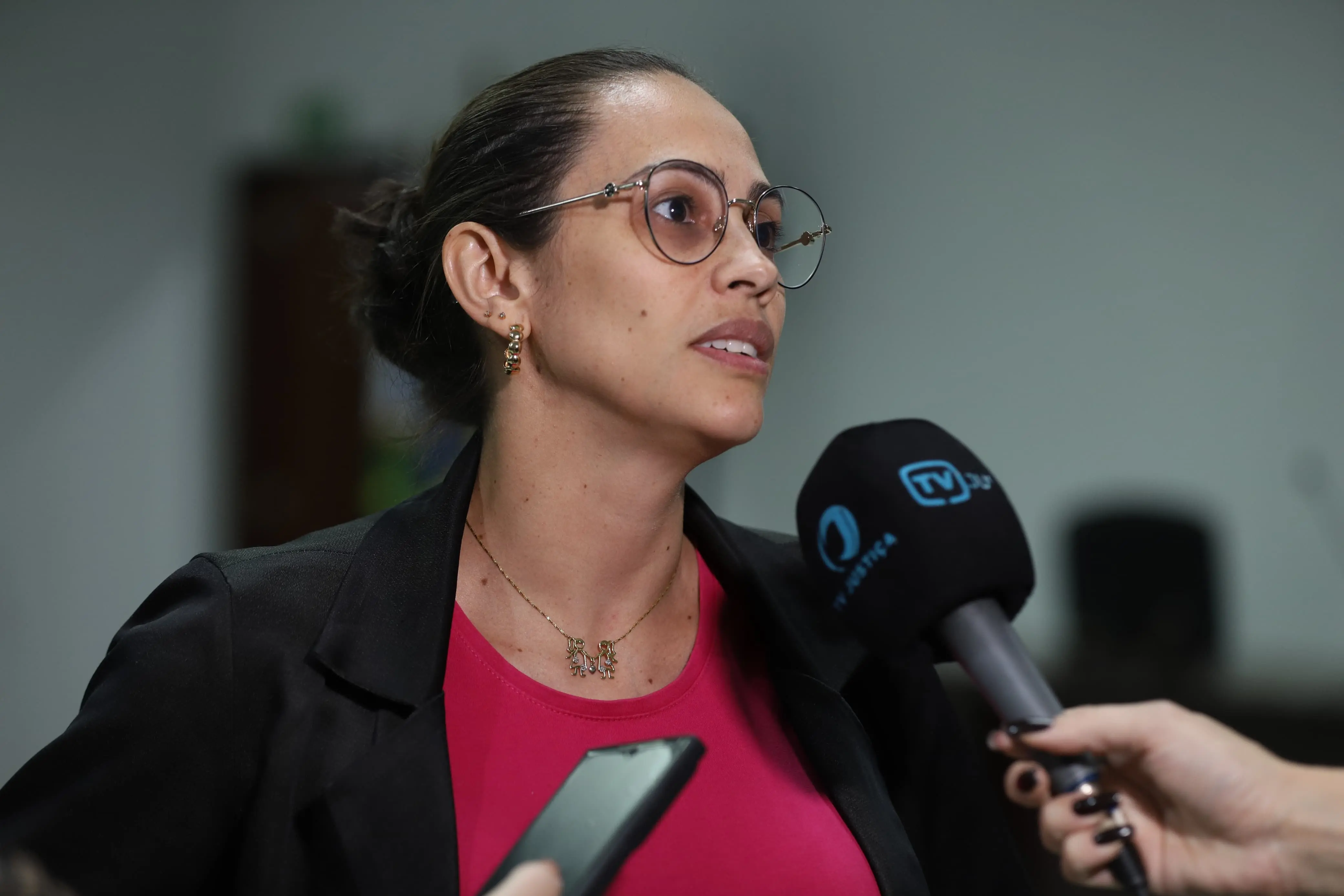 Mulher usa óculos redondos e blusa pink, com blazer escuro. Segura um microfone em entrevista. Promovendo acessibilidade e informação para todos.
