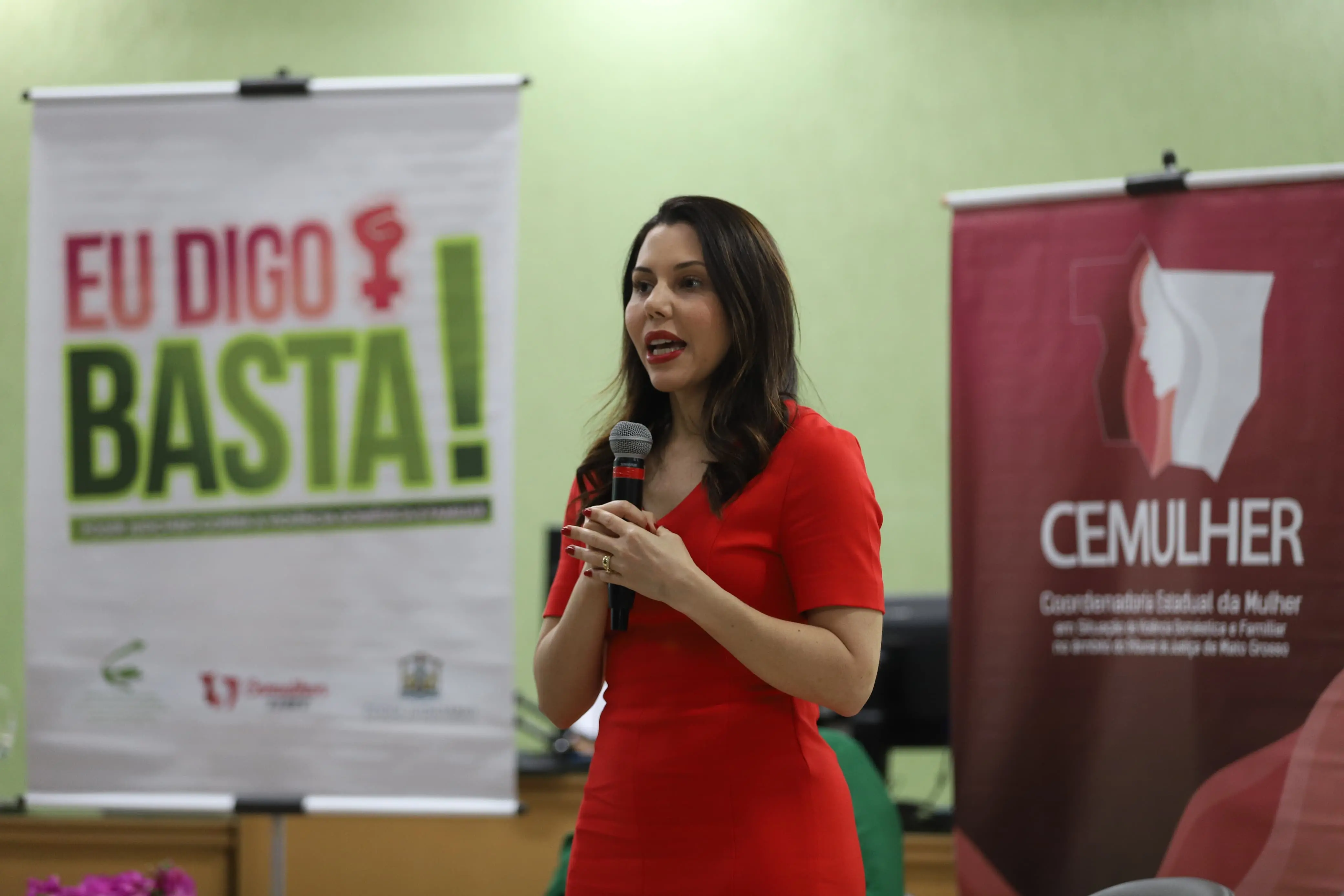 Mulher de vestido vermelho fala ao microfone com expressão séria. Atrás, banner da campanha “Eu Digo Basta!” e da Coordenadoria Estadual da Mulher (CEMULHER).