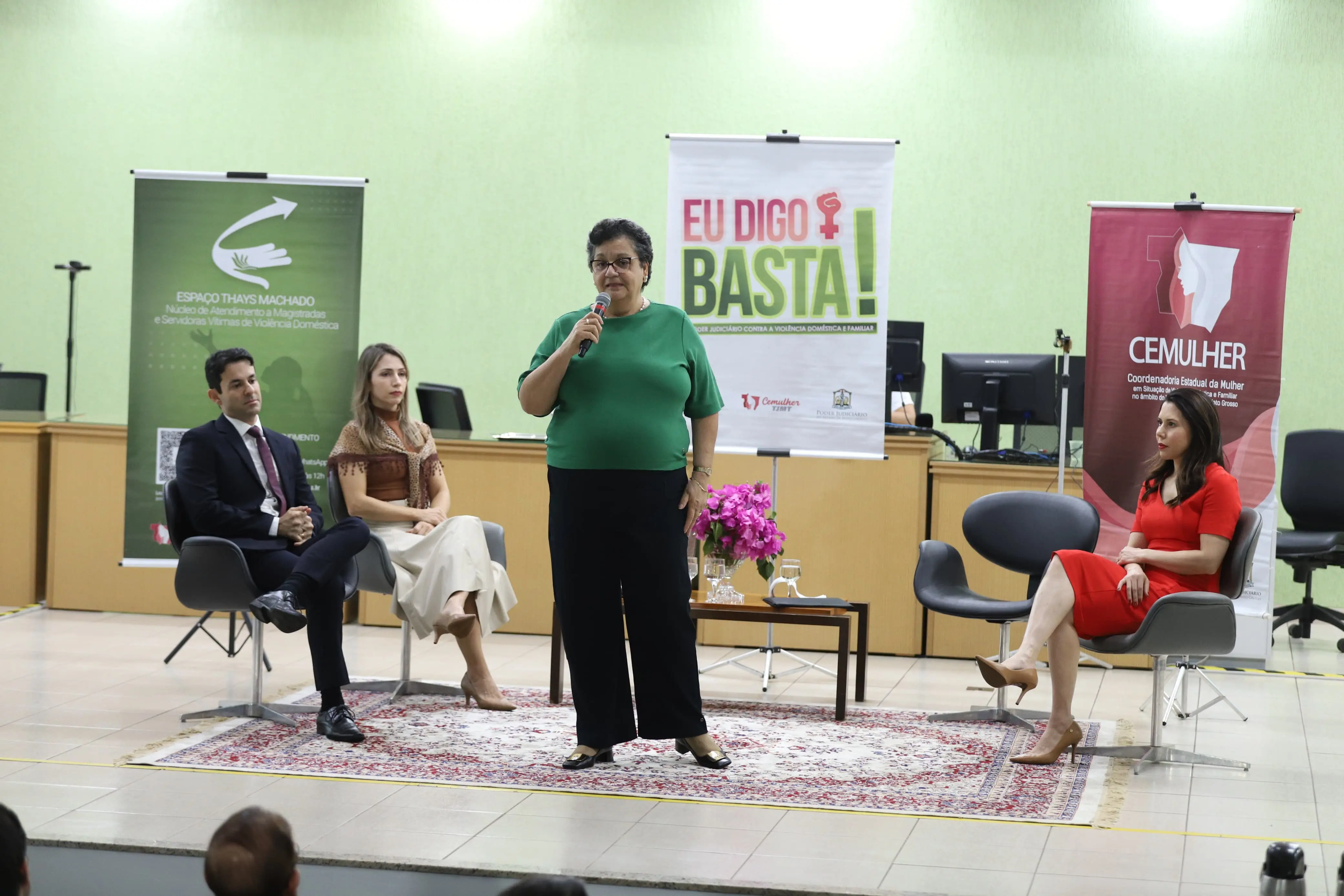 Mulher de blusa verde fala ao microfone no centro do palco. Outras três pessoas observam sentadas. Ao fundo, banners da campanha e decoração institucional.
