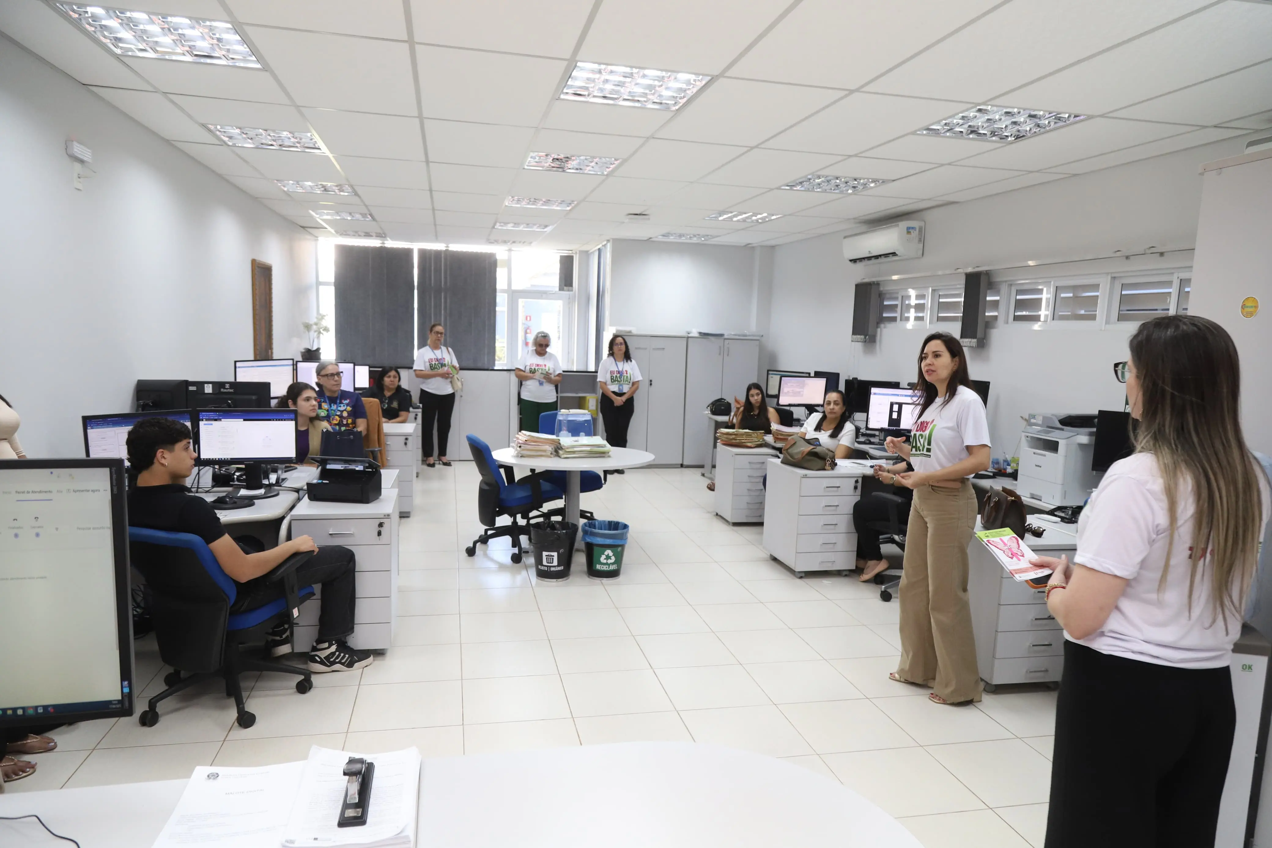 Ambiente de escritório espaçoso com várias pessoas trabalhando em computadores. Duas mulheres à direita, de camisetas "Eu Digo Basta!", conversam, enquanto outras circulam