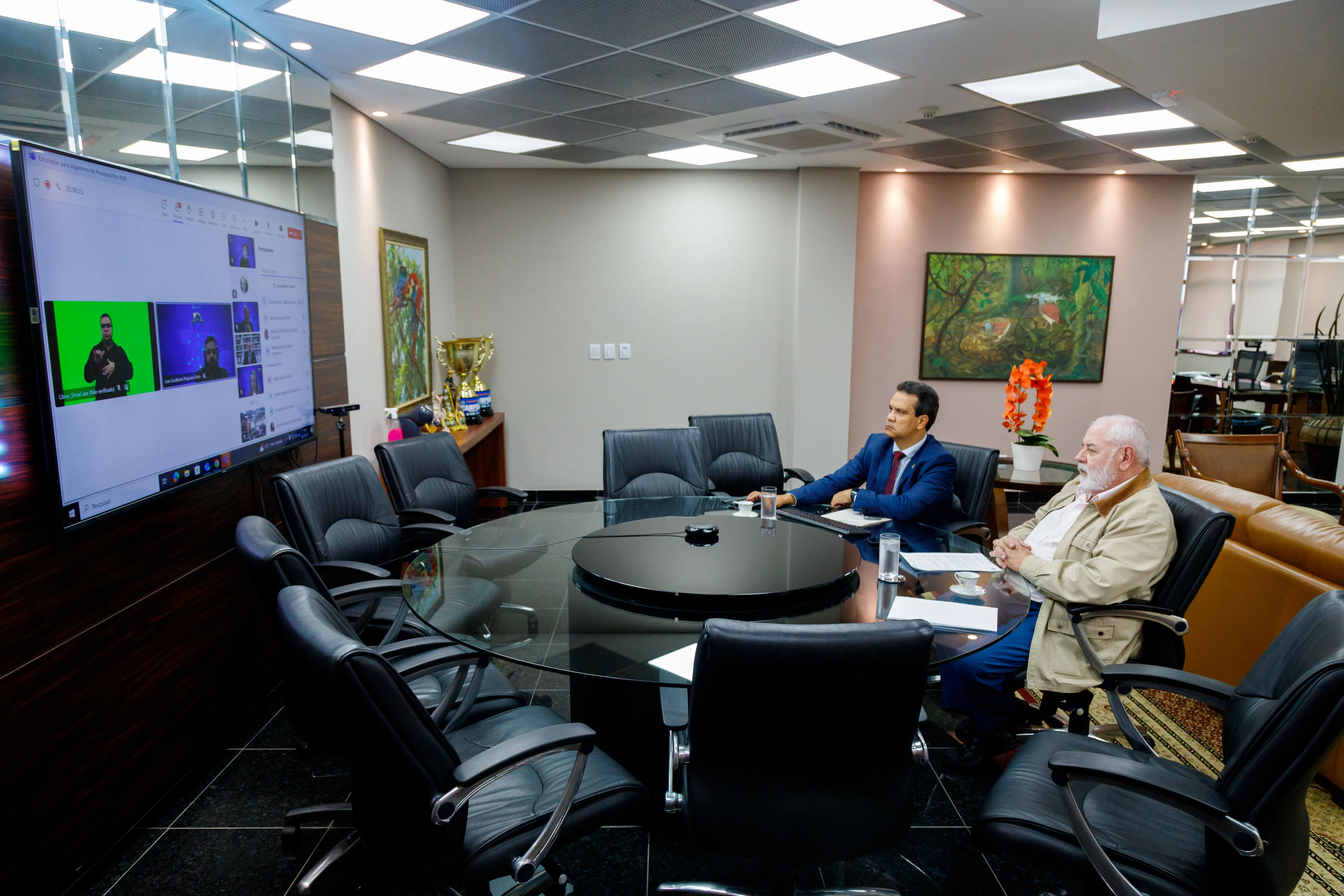 Presidente do TJMT e outro participante acompanham reunião virtual em sala ampla, com mesa redonda e cadeiras vazias. Na tela, aparece videoconferência com intérprete de Libras e demais participantes.